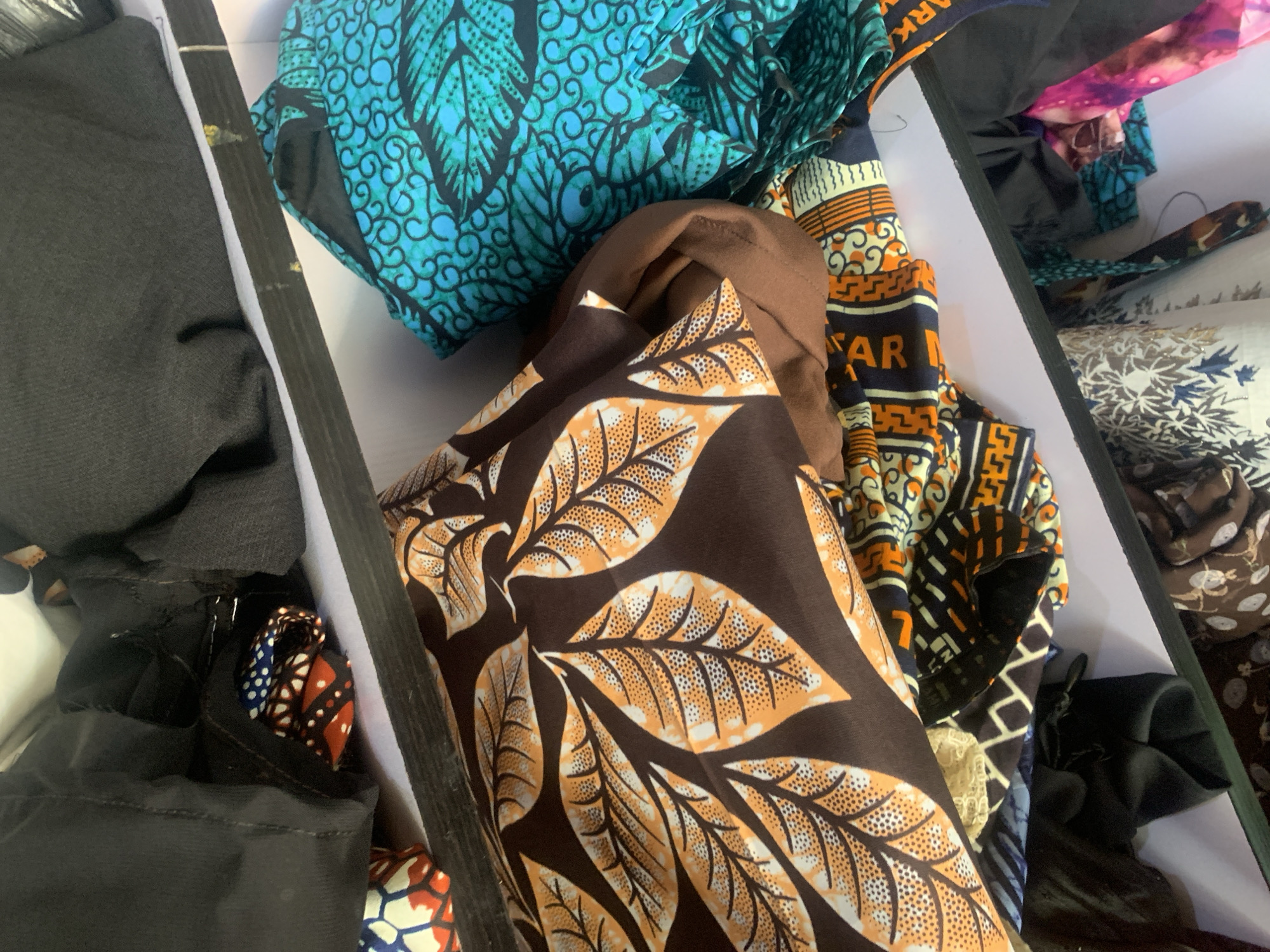 The image shows a close-up view of a shelf filled with fabric, likely in a market stall in Jauro Yinu, Nigeria. The fabrics are colorful and patterned, with some featuring large leaf motifs, geometric designs, and abstract swirls. Several black garments are also visible, draped over or tucked between the fabrics. The lighting suggests an indoor setting, possibly a market or shop. There are no people or animals present in the image.