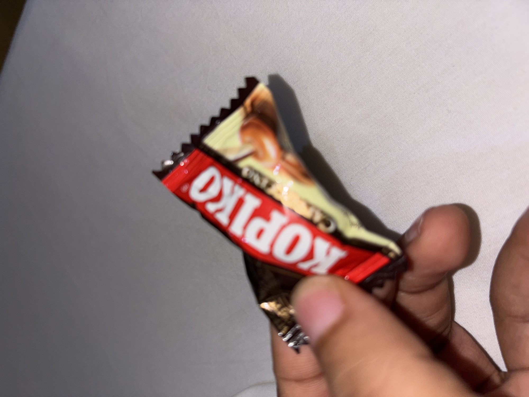 A hand holds a Kopiko candy wrapper against a light-colored fabric backdrop. The wrapper is red, yellow, and brown.