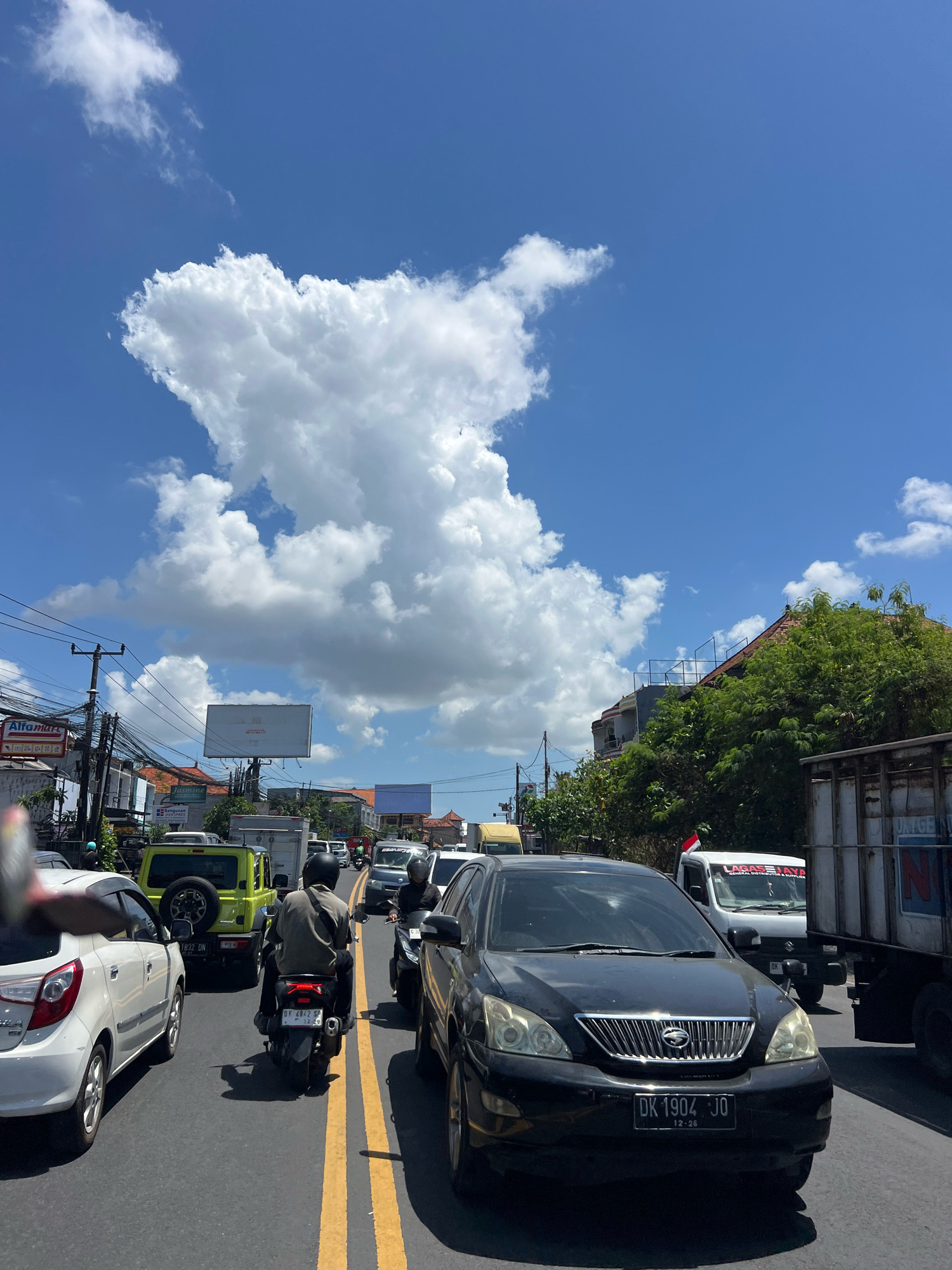 The image captures a traffic jam on a sunny day in Denpasar, Indonesia. The sky is a vibrant blue with large, fluffy white clouds. The foreground is dominated by a black car with license plate "DK 1904 JO". To its left is a white car and further ahead, a yellow SUV. A person on a motorcycle is visible in the middle lane, facing away from the camera. In the background, buildings, trees, power lines, and advertisements, including an "Alfamart" sign, are discernible. Several other vehicles, including trucks and cars, are part of the congested traffic. The presence of the license plate format and the "Alfamart" store chain indicates the location is likely within Denpasar, Bali, Indonesia, a popular tourist destination known for its traffic.