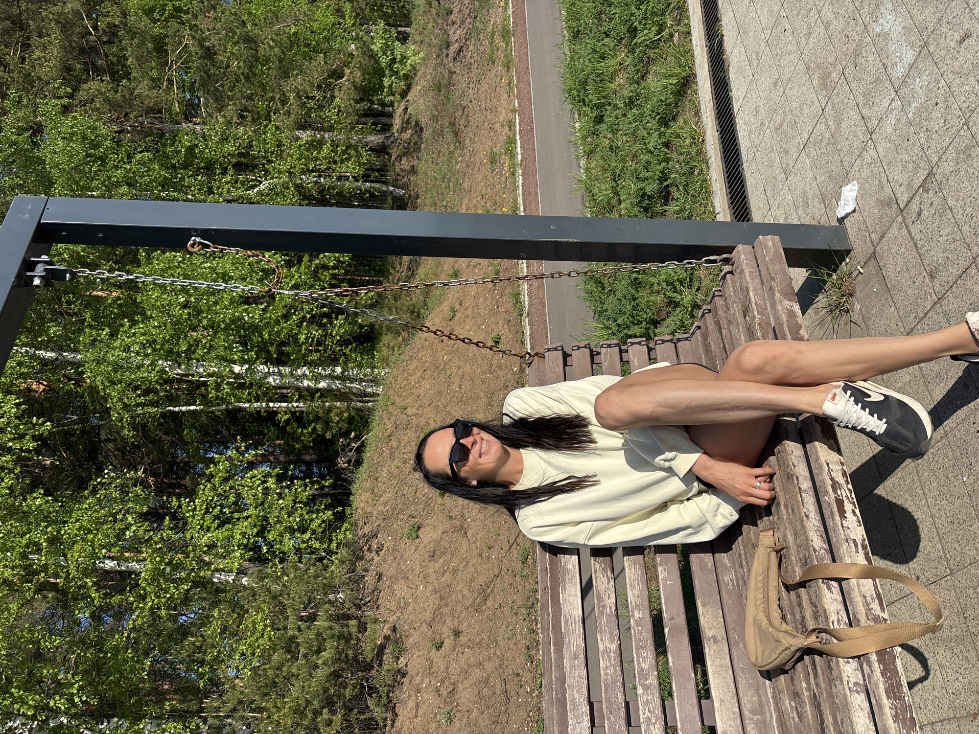 A woman with dark hair and sunglasses is lounging on a wooden swing bench. She is wearing a light-colored sweater and dark sneakers. Her legs are crossed at the ankles. A light brown bag rests on the bench near her feet. The bench is suspended by metal chains from a dark gray metal frame. Behind the bench is a dirt embankment and trees. To the right of the bench is a paved path. The location is Chaikovsky, Russia, as it is the city in which the image was taken.