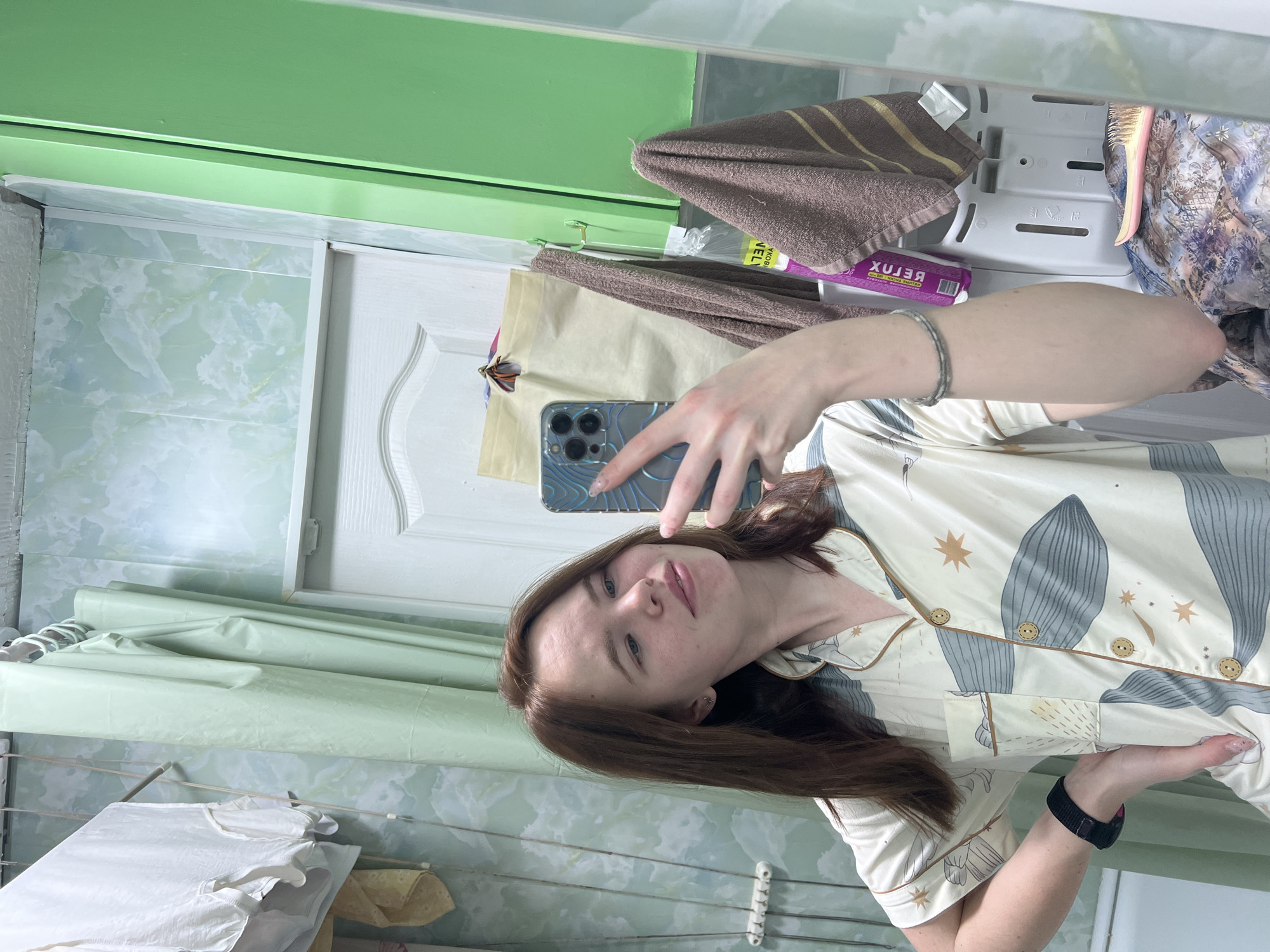 A woman is taking a selfie in a bathroom. She is wearing pajamas with a leaf and star pattern. She is holding a smartphone in her left hand, which is visible in the reflection of the mirror. She has brown hair and is wearing a black wristband on her right wrist and a bracelet on her left.
The bathroom has light green and white marble-patterned tile walls. A white door is visible. There is a clothesline with some clothes drying. Various items are stored on a rack above the door, including towels and a box of "RELJAX" medication.
It's impossible to pinpoint the exact location in Chaikovsky, Russia, without further information.