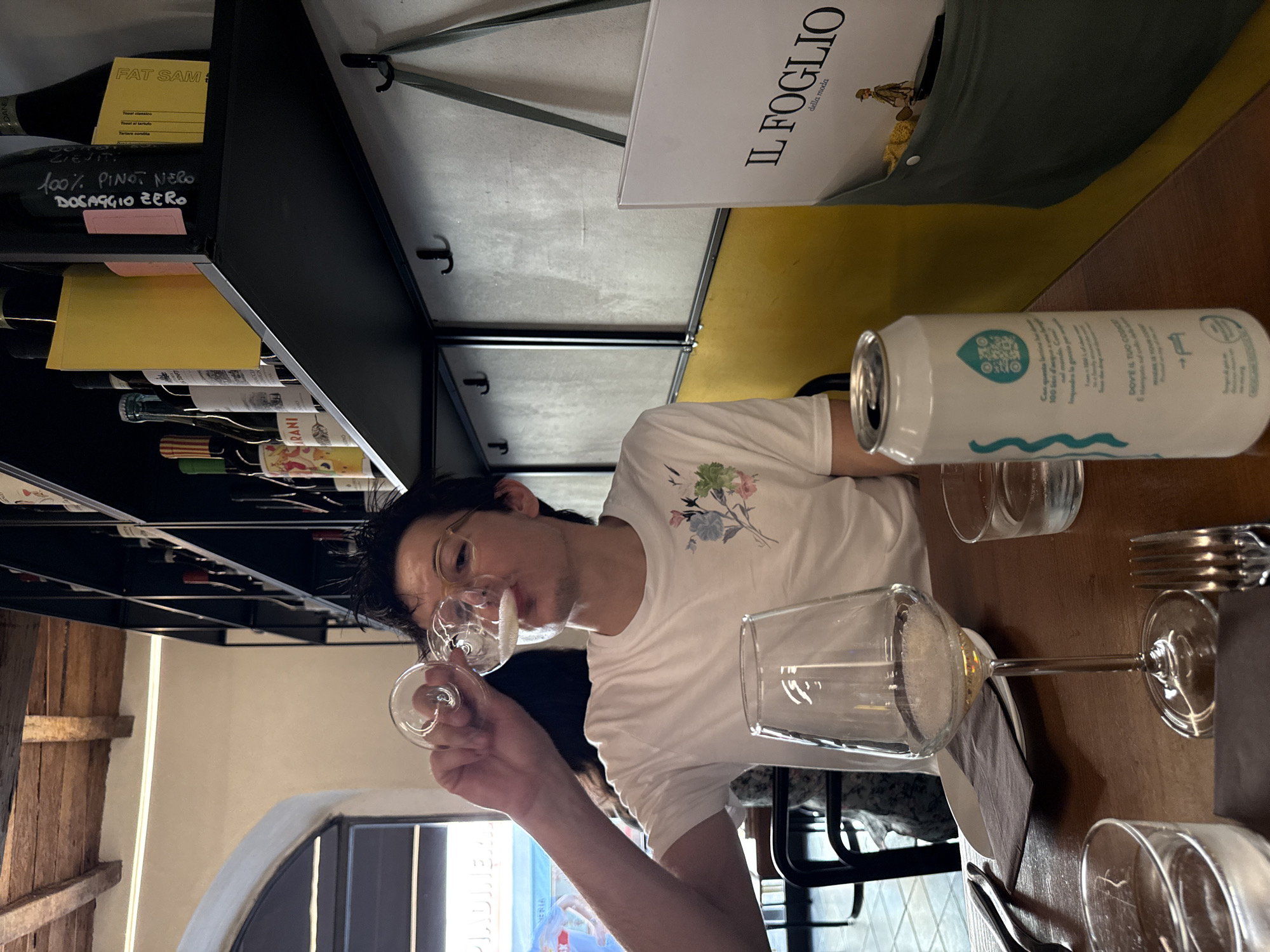 Here is a description of the image:
The image depicts a woman tasting wine at a table, likely in a restaurant in Milan, Italy. She is wearing glasses and a white t-shirt and holds a small wine glass to her lips. Several other glasses are on the table, along with cutlery, plates, and a can of sparkling water. A wine rack is visible in the background, displaying various wine bottles with handwritten labels. A menu or sign with the text "IL FOGLIO della moda" is also present.