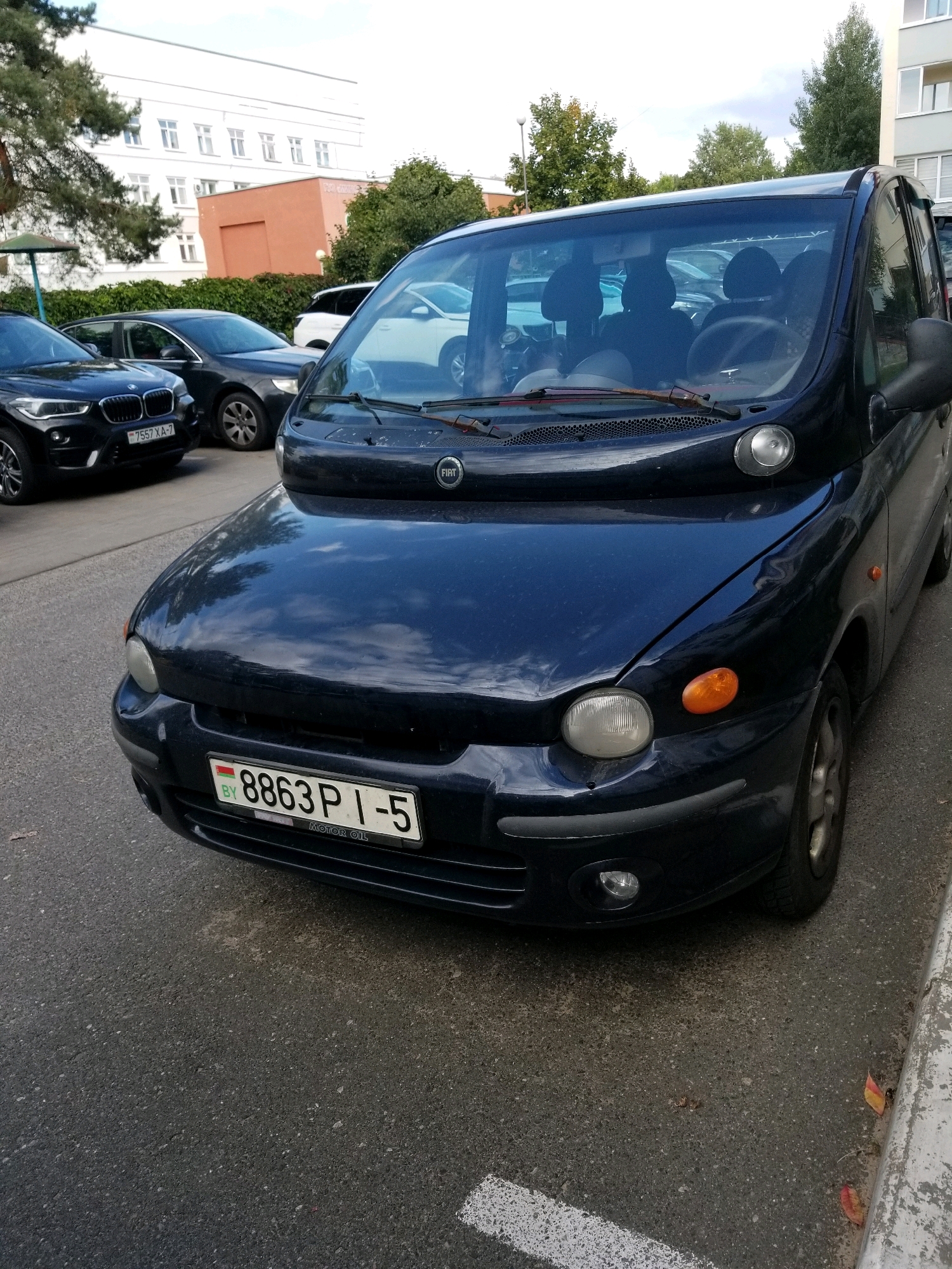 The image shows a dark blue Fiat Multipla parked on an asphalt surface, likely a street or parking lot, in a city setting. The car's license plate is visible and reads "8863 P I-5 BY". In the background, there is a large white building with multiple windows, suggesting an urban or institutional area. Trees and greenery are also present, indicating an outdoor environment. The car is the central focus of the image, and its front end, including the headlights and grille, is clearly visible. The overall scene appears to be a daytime shot under overcast or partly cloudy conditions. The location is identified as Бараўлянскі сельскі Савет, Belarus.