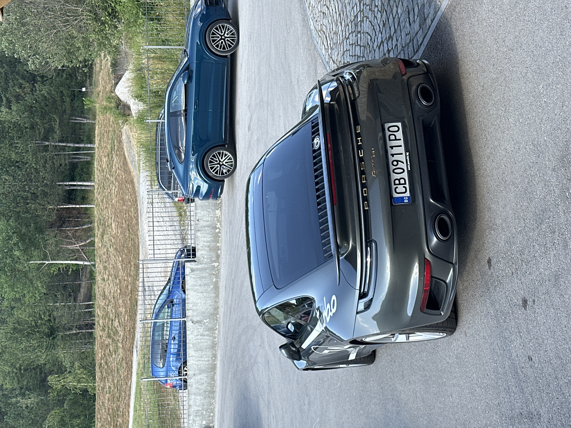 In Kerenite, Bulgaria, three cars are parked on asphalt and cobblestone surfaces, with a wooded hillside in the background. In the foreground, a dark gray Porsche 911 Turbo is parked at an angle, facing away from the viewer. The car has a prominent rear spoiler, dual exhaust pipes, and a license plate reading "CB 0911 PO." A white stripe with the number "14" is visible on the rear fender. To the left, a blue Volkswagen Golf is parked behind a metal fence and concrete barrier. Further back and to the right, a dark teal Porsche Cayenne Coupe is parked. The environment is outdoors, with vegetation and a clear sky.