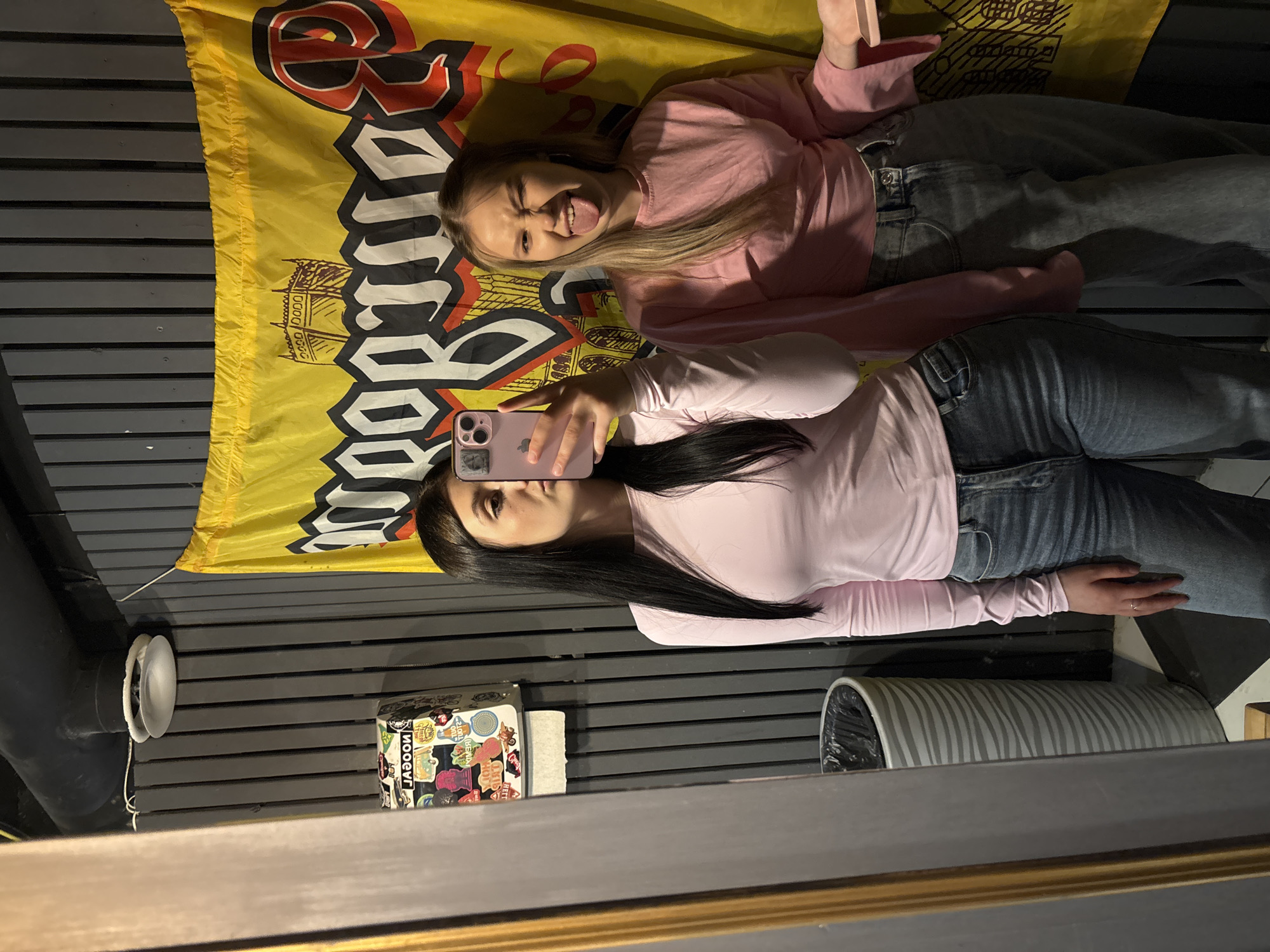 The image depicts two young women within an indoor establishment located in Chaikovsky, Russia. The environment features dark grey vertically paneled walls, a partial view of a black and white checkered floor, and exposed dark pipes near the ceiling.
On the left side, a woman with long, dark hair, wearing a light pink long-sleeved top and blue jeans, is reflected in a rectangular mirror with a gold frame. She holds a pink smartphone to her face, appearing to take a mirror selfie, with her lips lightly puckered. A silver ring is visible on her left ring finger. Within the mirror's reflection, to her left, stands a tall, cylindrical trash can with a grey and white wavy pattern and a black liner. Above the trash can, a white dispenser or box, heavily adorned with colorful stickers, is mounted on the dark grey wall, with a roll of white paper positioned beneath it.
To the right of the first woman, a second woman with blonde hair, dressed in a loose-fitting pink long-sleeved top and grey jeans, stands making a playful expression. She has her tongue extended and one eye squinted. Behind both women, a large yellow banner or flag hangs prominently on the dark grey wall. This banner displays black and red stylized text, including visible elements resembling "GORN" or "GORNY" and a large "R," along with an illustration of a castle-like structure. A round, frosted glass light fixture is mounted high on the dark grey wall above the banner.