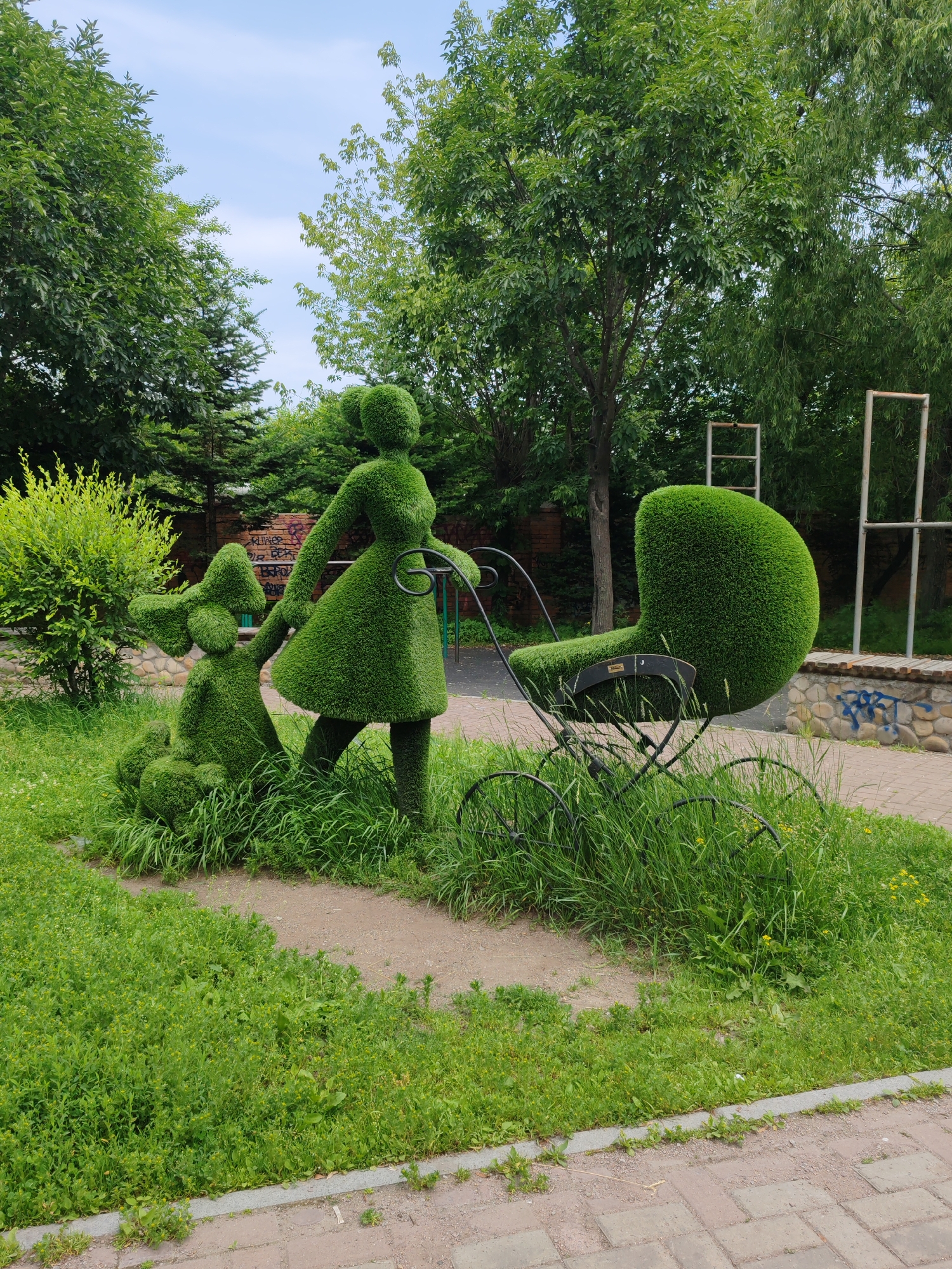 The image displays a topiary sculpture of a woman and a dog next to a baby carriage. The sculptures are meticulously crafted from green shrubs, resembling the figures in motion. The woman is depicted in a dress, holding the dog's leash, while the dog sits beside her. The baby carriage, also made of topiary, is positioned to the right of the woman.
The sculptures are situated in a park-like setting with lush green grass and trees in the background. A paved walkway runs through the scene, suggesting it's a public area. The sky is visible above, with scattered clouds. The presence of graffiti on a low brick wall in the background indicates an urban or semi-urban environment.
The location is a park in Vladivostok, Russia. The topiary art is a prominent feature of the landscape, likely a tourist attraction or a decorative element in a public space. The overall atmosphere is serene and picturesque, with the vibrant green of the sculptures contrasting with the natural surroundings.