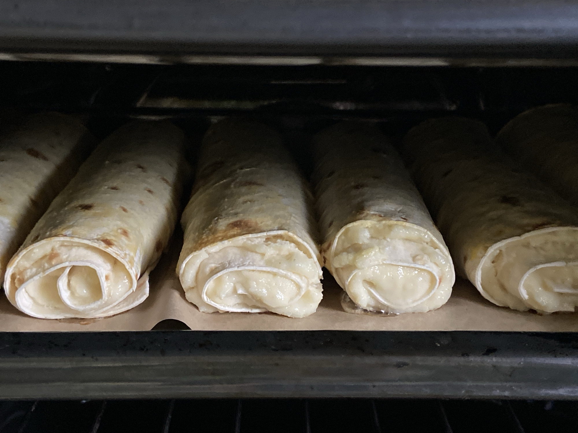 The image displays a close-up, eye-level view of six cylindrical, rolled food items, likely tortillas or crepes filled with a creamy-white substance, baking inside an oven. The food items are arranged in a single row on a sheet of light brown parchment paper, which rests on a dark metal rack or baking tray.
The filling in each roll appears melted and slightly oozing or bubbling from the ends, suggesting it could be cheese or a sweet cream that has been heated. The wraps themselves show areas of light golden-brown color, indicating they are in the process of cooking or have recently been cooked. The interior of the oven is dark, with shadowy areas above and behind the food, and some reflections on the dark metal surfaces. The scene captures the final stages of preparing these rolled treats in a domestic kitchen setting. There are no people visible, and no text or specific location cues beyond the oven itself. The overall impression is of food being actively prepared.