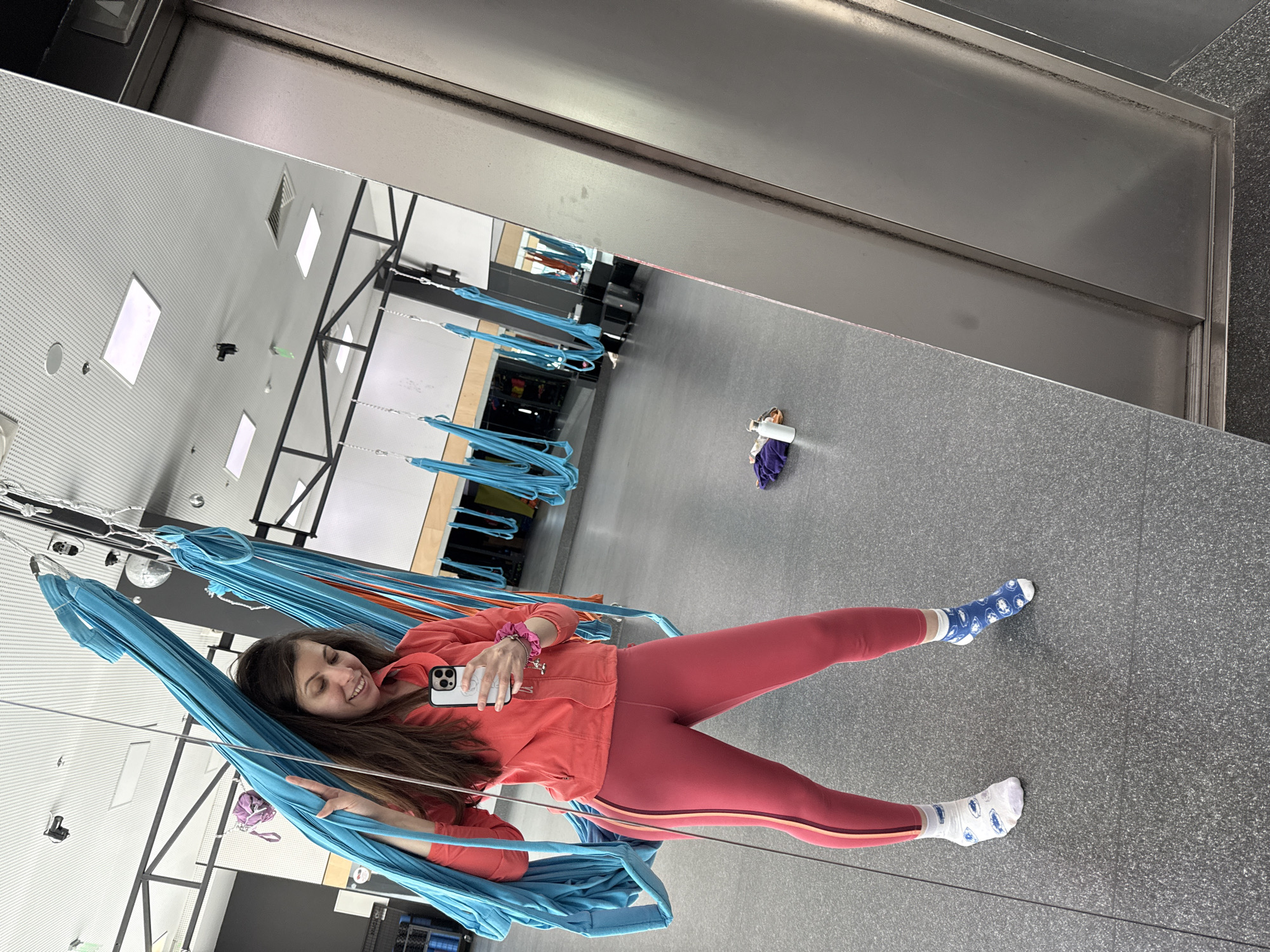 A woman in pink leggings and a coral jacket is reclining in a blue aerial hammock, holding a phone in her hand. She is smiling and appears to be taking a selfie. The hammock is suspended from a black metal frame. In the background, more aerial hammocks are visible. The floor is gray. The room appears to be a studio for aerial yoga or similar activities. A bag is lying on the floor to the right of the woman.