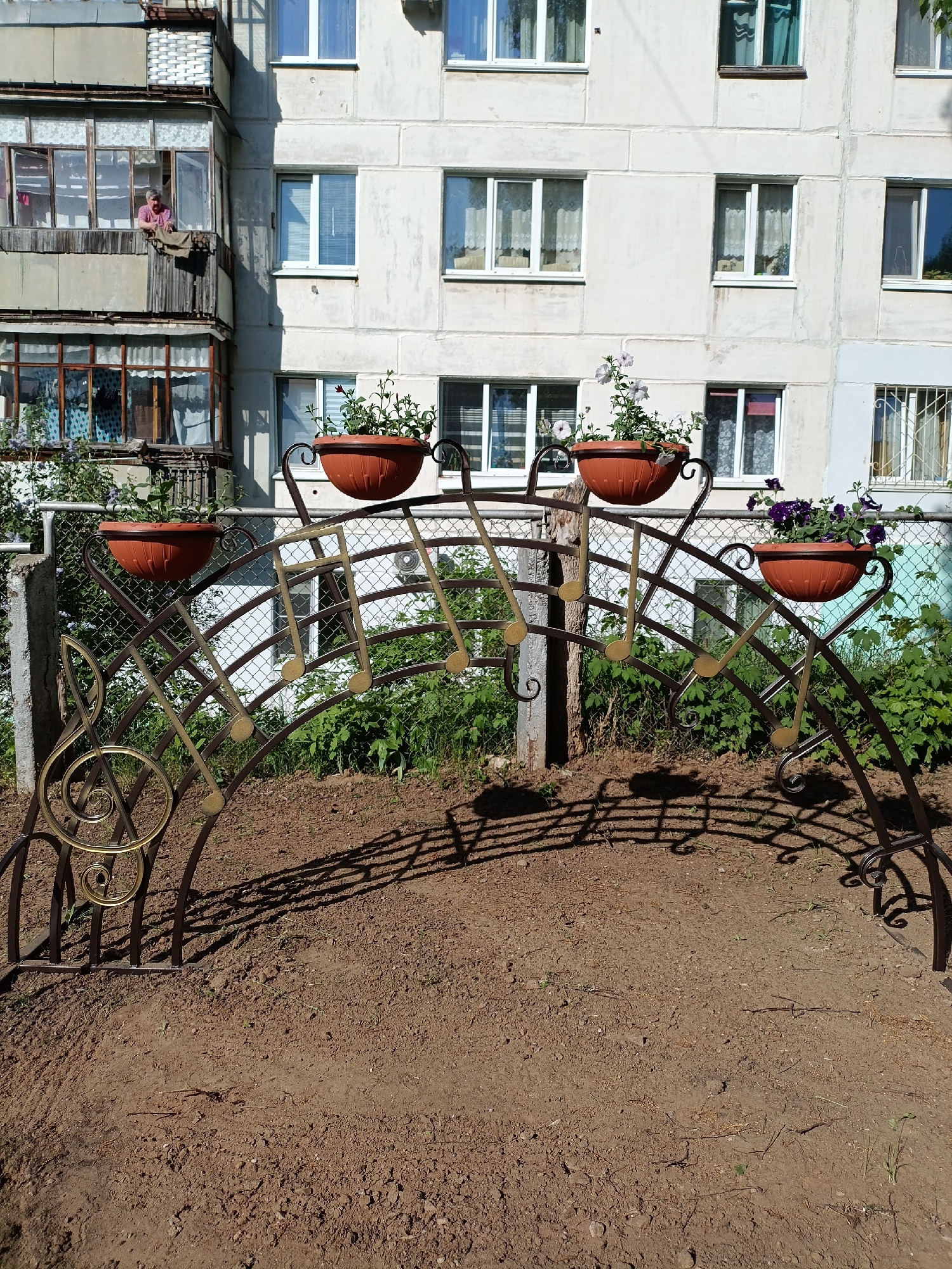 The image depicts a metal archway with musical note designs supporting flower pots, set in a patch of bare earth. The archway, constructed of dark metal with golden accents on the musical notes and decorative swirls, stands in front of a chain-link fence partially obscured by greenery. Four terracotta-colored flower pots are positioned atop the arch, containing various flowering plants.
In the background, a multi-story residential building dominates the scene. The building has a façade of light-colored concrete, with a series of rectangular windows arranged in a grid pattern. On the left side of the building, a balcony is visible where a person is standing looking out over the balcony.
The scene is located in the city of Chaikovsky, Russia.