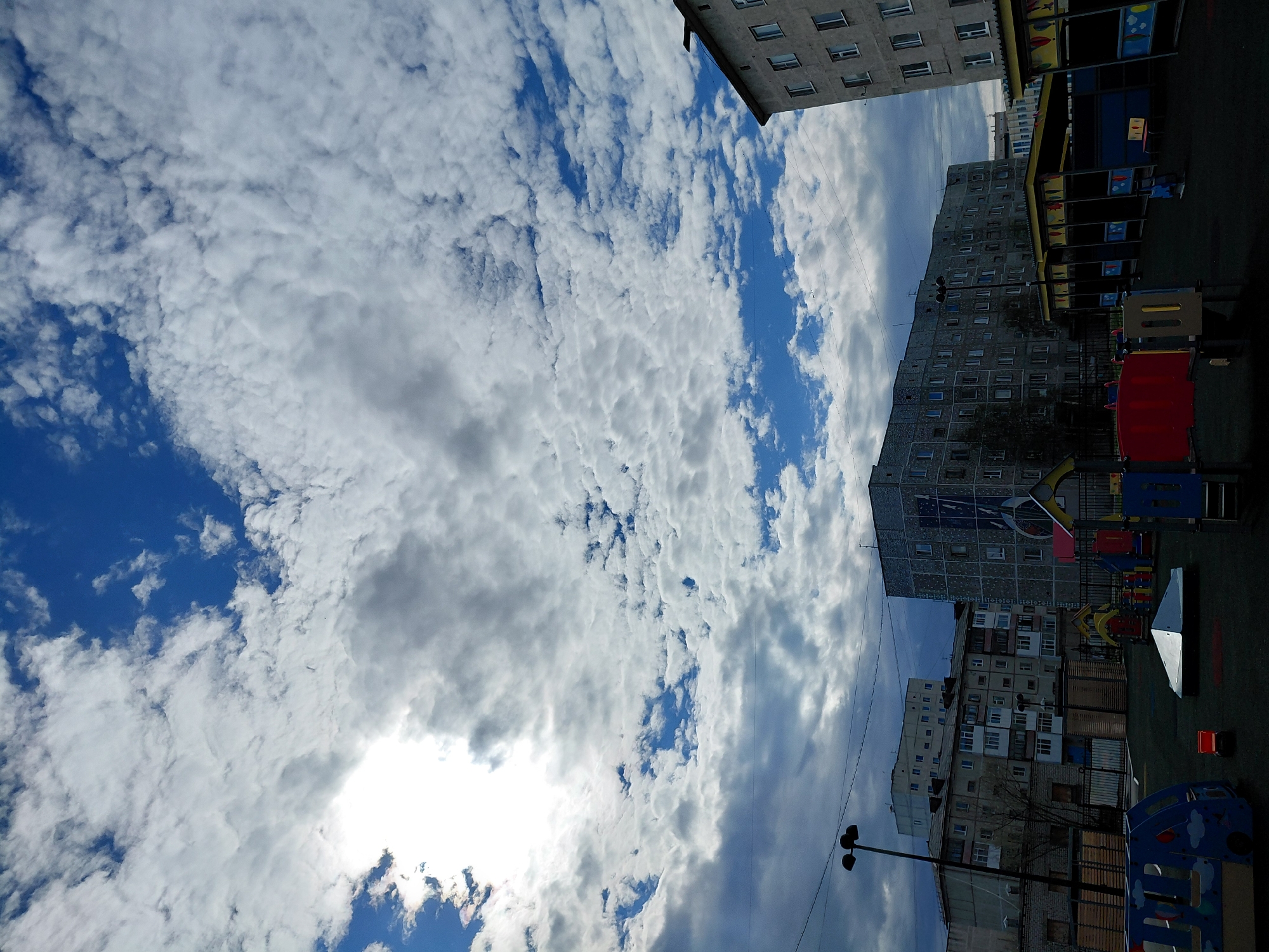The image captures an urban residential scene in Nadym, Russia, under a dynamic sky. The sky dominates the upper and central portions of the photograph, featuring bright blue patches interspersed with a wide array of white and grey clouds, suggestive of a partly cloudy day. A bright area on the left hints at the sun's location, partially obscured by clouds.
In the lower and right parts of the frame, several multi-story apartment buildings are visible. These structures appear to be concrete or stone, with numerous windows. One prominent dark grey building in the mid-right features a large mural or artistic design on its side, facing the viewer. Adjacent to and in front of these buildings is a children's playground, equipped with colorful structures in red, blue, and yellow, likely including slides or climbing frames. The ground of the playground appears to be a dark, possibly artificial surface.
Additional objects include a black streetlight pole with multiple lamps positioned on the left side, and several overhead utility wires stretching across the sky, connecting to the buildings. The overall setting depicts a typical daytime scene in a residential district, characterized by Soviet-era architectural influences common in Russian cities. No people or discernible text are visible in the image.