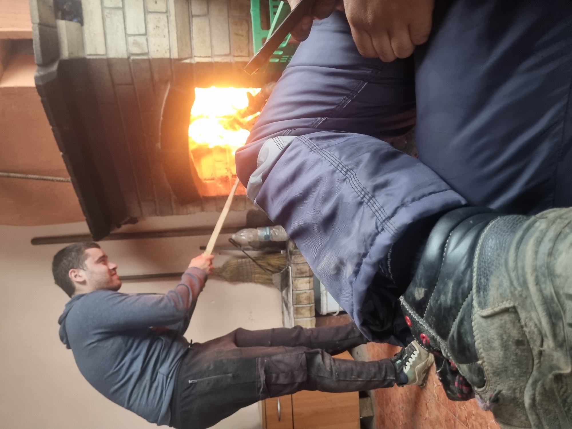 A man is using a long, thin stick to tend a fire in a brick oven. The oven is in a room with a light-colored wall, a broom, and a cabinet. Another person is in the foreground, partially obscured, wearing dark pants and boots. The location is Ovcharovo, Bulgaria.