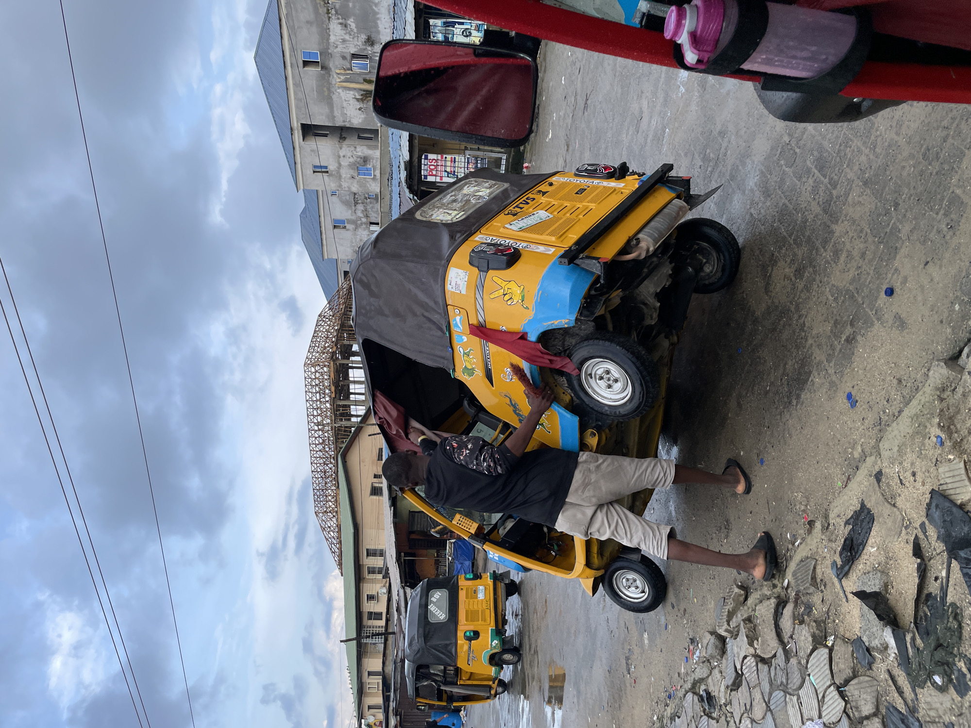 An urban street scene in Ado, Nigeria, is depicted under an overcast sky. The ground is wet and appears muddy in sections.
In the center, a dark-skinned man in a black long-sleeved shirt with patterned sleeves and light-colored shorts, wearing black sandals, is attending to a yellow and blue tricycle (keke napep) that is tilted onto its right side. The man's hands are positioned within the engine compartment or under the passenger seat, suggesting maintenance or repair. The tricycle features "TVS" and "TOYOTA" branding, a "DELUXE" sticker, a red fabric strip tied to the rear, a yellow peace sign decal, and a lion head graphic.
Further down the street to the left, a second yellow tricycle with a black canopy is parked, displaying "KEREKE BOY" text on its rear.
In the immediate foreground on the right, a large red side mirror is partially visible, and a pink water bottle secured in a black holder is attached to a red vertical bar.
The background features various buildings. To the left, a light-colored building with a green roof is visible. In the central background, a significant construction project with an exposed, intricate wooden or metal roof framework stands incomplete. To the right, multiple gray concrete buildings with blue metallic roofs and rectangular windows, some appearing unfinished, line the street.
Overhead, several dark power lines stretch horizontally across the cloudy sky. The foreground on the left includes a broken and uneven paved sidewalk littered with debris.