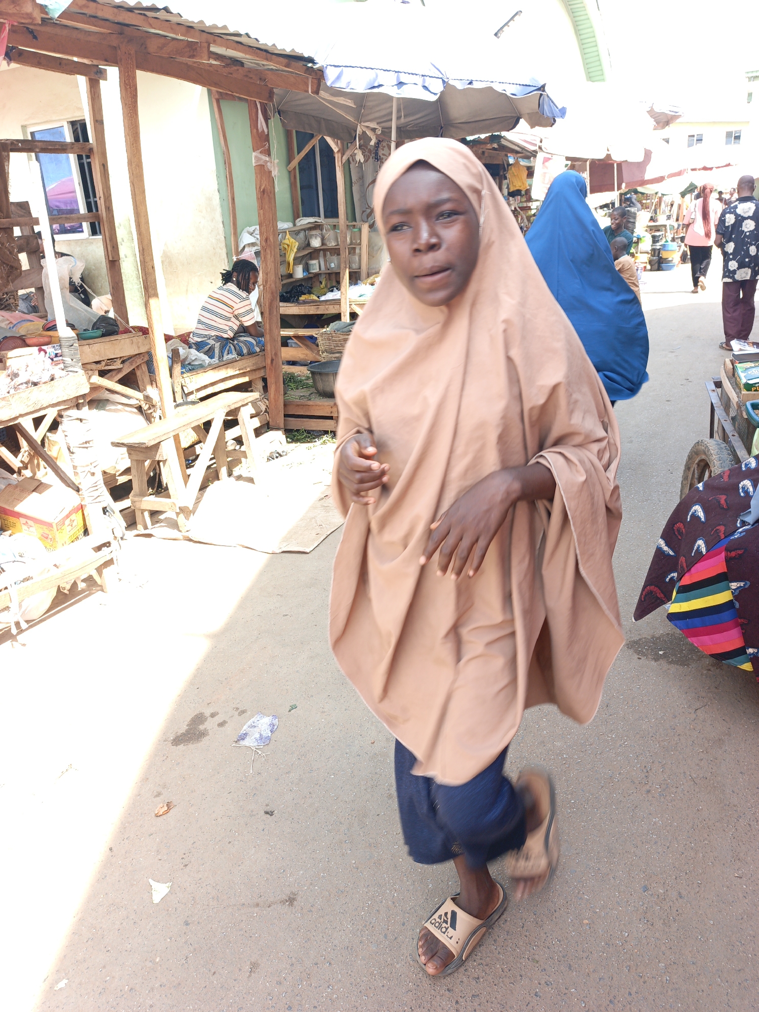 A young person in a light-brown hijab and blue pants walks in the foreground of a street scene in Akwanga, Nigeria. The individual wears tan Adidas sandals. Behind them are market stalls under makeshift canopies, displaying goods and food. Other people are walking in the background. Trash is scattered on the road's surface. The location appears to be a bustling outdoor market.