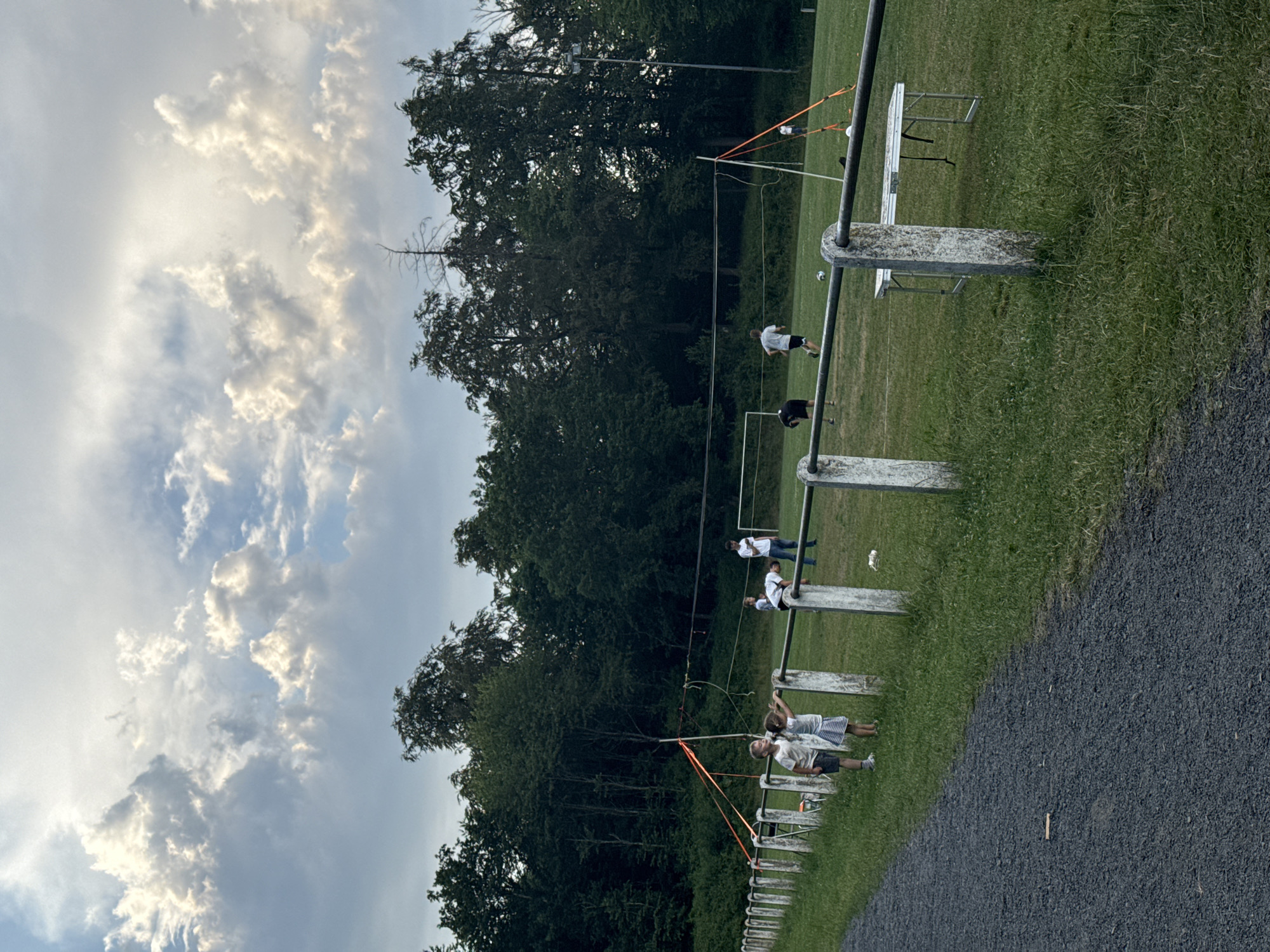 The image depicts an outdoor scene on a grassy field, likely a sports ground, with a line of white posts and a metal railing in the foreground, separating a gravel path from the field. In the background, a dense line of trees forms the edge of the field, and beyond that, more trees can be seen. The sky above is filled with dramatic clouds, catching the light.
Several individuals are present on the field. In the left foreground, two children, a boy and a girl, are standing by the railing, looking towards the field. Further back on the field, a group of people appear to be engaged in some activity, possibly a game or practice. One person is bent over near the railing on the right side of the field. A volleyball net is set up across the field, with orange straps visible supporting the net posts. A folding table is also present on the field. A white soccer goal is visible in the distance. The setting appears to be Lumda, Germany, given the provided context.