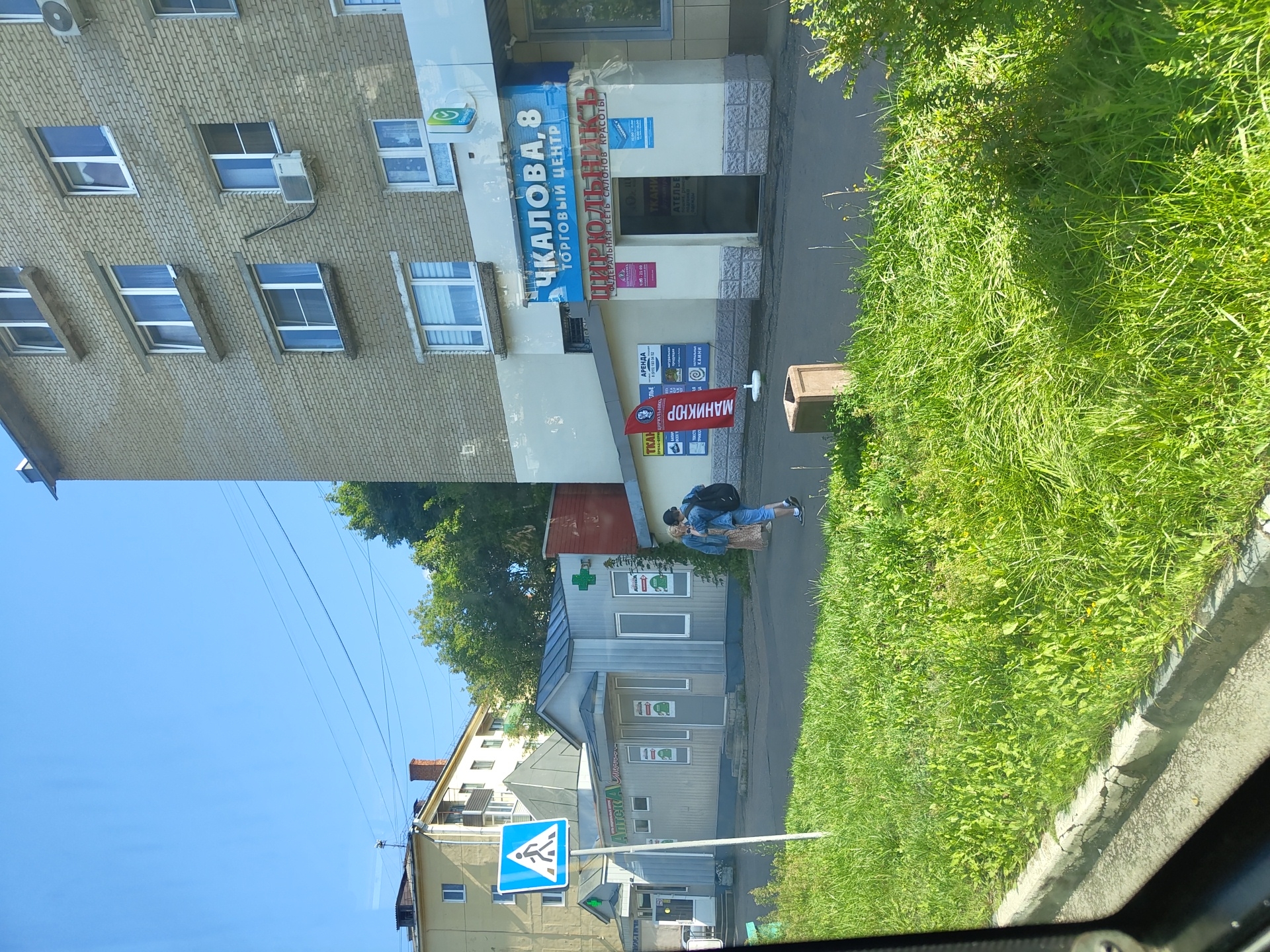 The image depicts a street scene in Zhukovsky, Russia, on a sunny day. In the foreground, a lush green grassy verge lines the paved sidewalk. A person wearing a blue denim jacket, light-colored pants, and a backpack is walking away from the camera on the sidewalk. They are walking towards two other individuals standing near a small, light-colored building with a gray roof. This building appears to be a pharmacy, indicated by a green cross symbol and the word "АПТЕКА" (Apteka - Pharmacy) on its facade.
To the right of the pharmacy, a larger, multi-story brick building stands prominently. A blue sign above an entrance reads "ЧКАЛОВА, 8 ТОРГОВЫЙ ЦЕНТР" (Chkalova, 8 Trading Center). Below this, another sign in red states "ПИРЮЛЬНИК" (Pirjul'nik - Hairdresser) with smaller text identifying it as a federal chain of beauty salons. Various advertisements and signs are visible on the facade of this building.
Further to the left, another building, predominantly yellow, is partially visible. A blue pedestrian crossing sign with a white silhouette of a person is positioned near the sidewalk. Power lines are visible stretching across the clear blue sky. The overall environment suggests a commercial or residential area within the city.