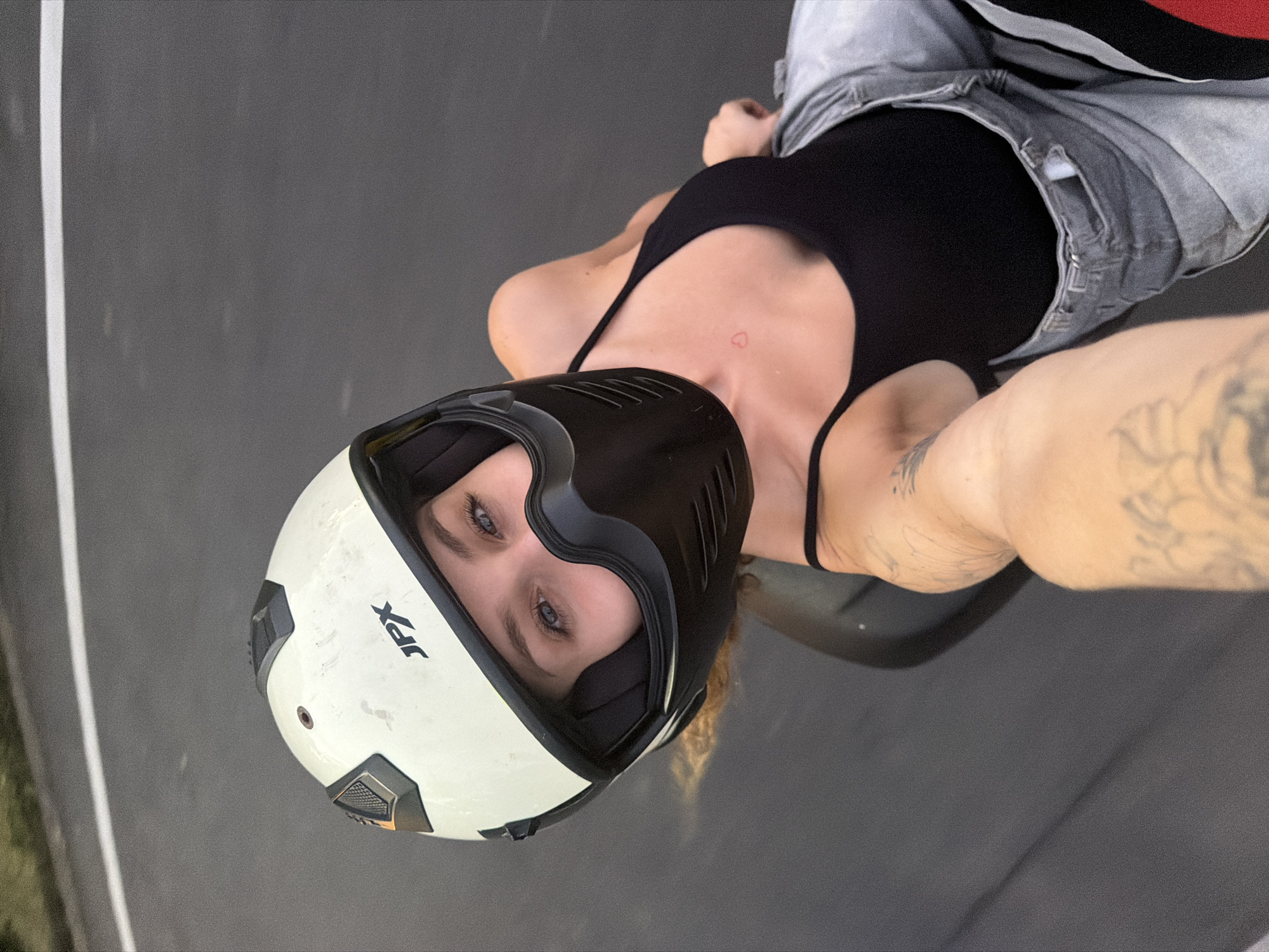 A woman wearing a white and black helmet, black tank top, and grey jeans, sits on a motorcycle. Her face, with blue eyes and light brown hair, is visible through the helmet's opening. She has a small red heart tattoo on her chest and tattoos on her arms. The motorcycle is on a grey asphalt road with a white line running across it. The background shows a blurred green area. The scene is likely Mengwi, Indonesia, based on the provided information.