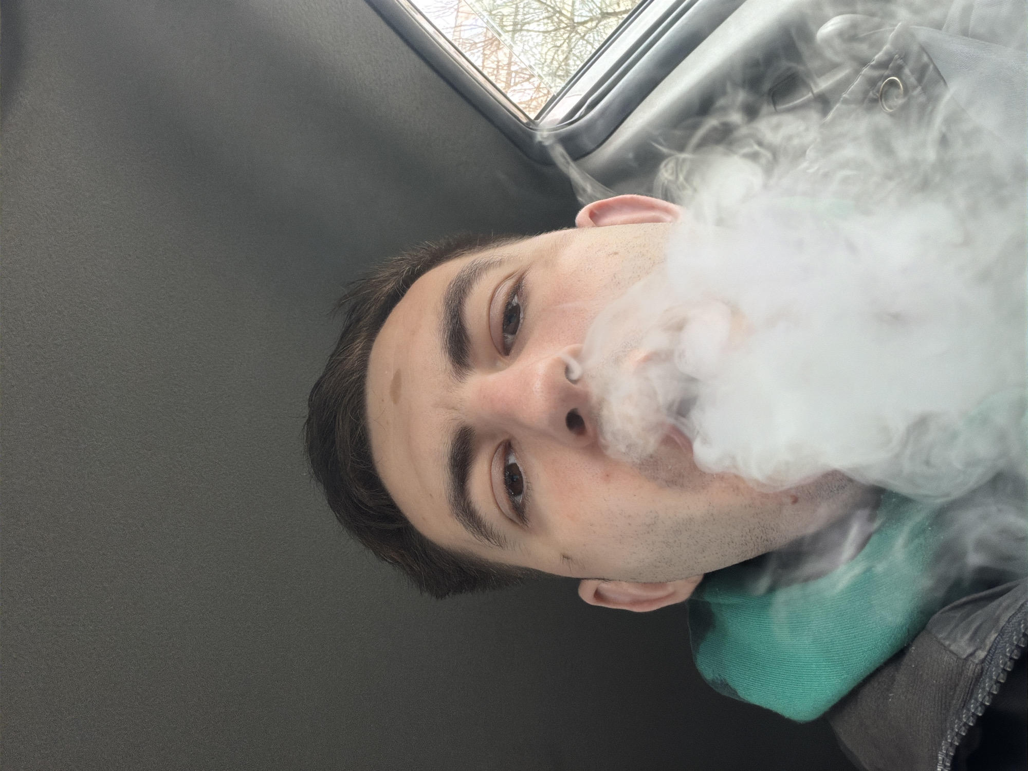 A close-up selfie shows a man exhaling a large cloud of white smoke. The man has dark hair, light skin, and brown eyes. He wears a green garment around his neck and a dark jacket or coat. The background shows a gray surface, possibly the interior of a vehicle, and a window with bare tree branches visible outside. The location is not discernible as Varna, Bulgaria, from the visual evidence.