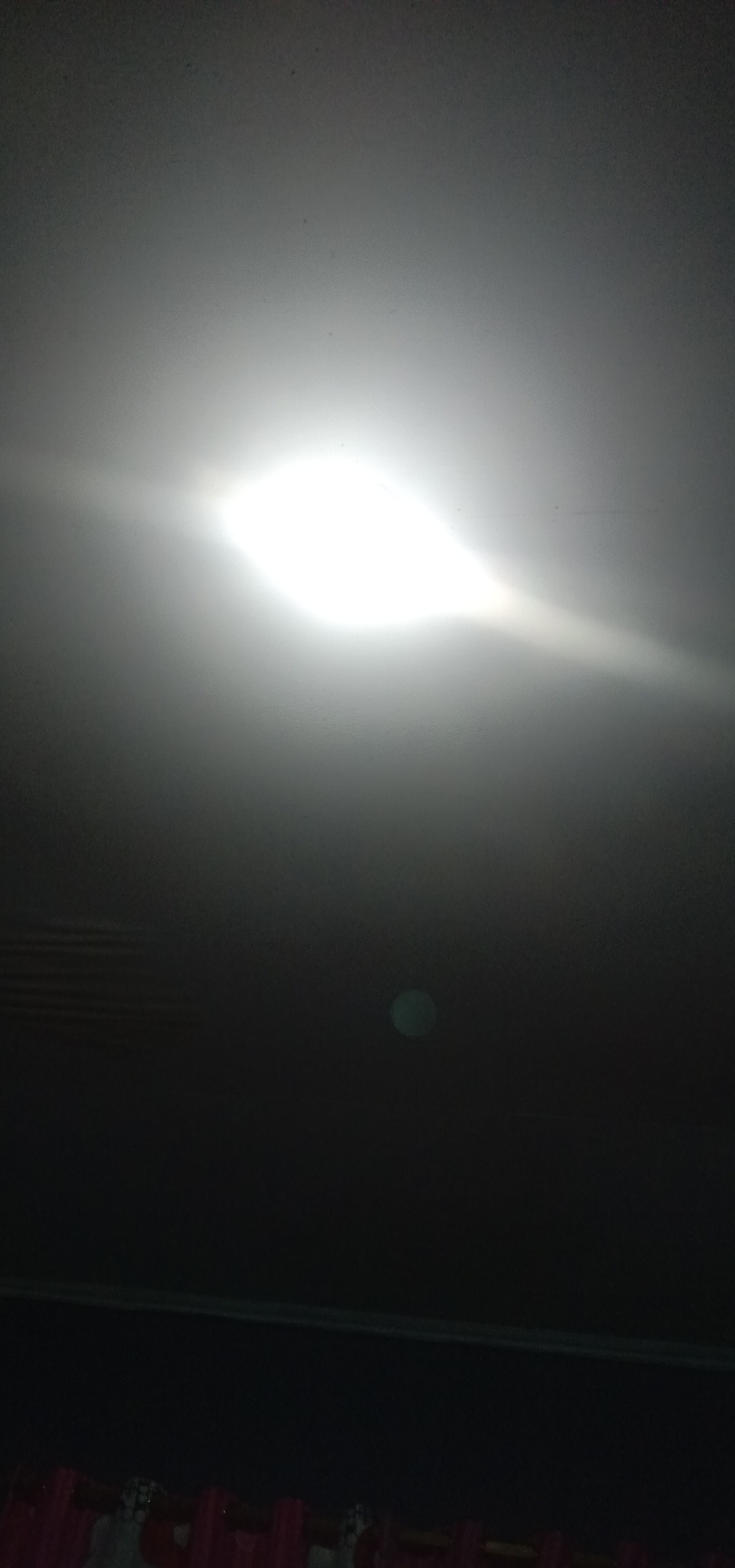 The image depicts a dark room with a bright light source in the upper half, potentially a ceiling light or a flash from a camera. The light creates a halo effect and illuminates the dust particles in the air, giving the scene a hazy appearance. The lower portion of the image is predominantly dark, with a horizontal line across the middle that might be a shelf or the edge of a piece of furniture. Below this line, the bottom of the image shows the tops of pink and white curtains. The overall impression is that of an indoor environment. There are no people or clear subjects visible in the image.