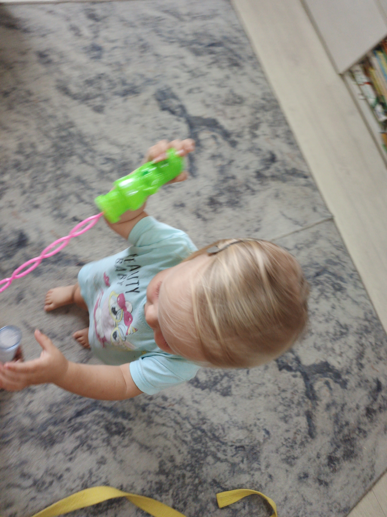 A young child with blonde hair is sitting on a carpeted floor and playing with a green bubble gun that has a pink string attached. The child is wearing a light blue t-shirt with a cartoon owl graphic. The child's left hand is holding a small, silver cylindrical object. The carpet is gray with a textured pattern. To the right of the carpet, there is a light-colored wooden floor and a shelf with books. The image appears to be taken indoors, likely in a residential setting, as there are no visible windows or outdoor elements. There is no indication of the city of Zlatoust, Russia.