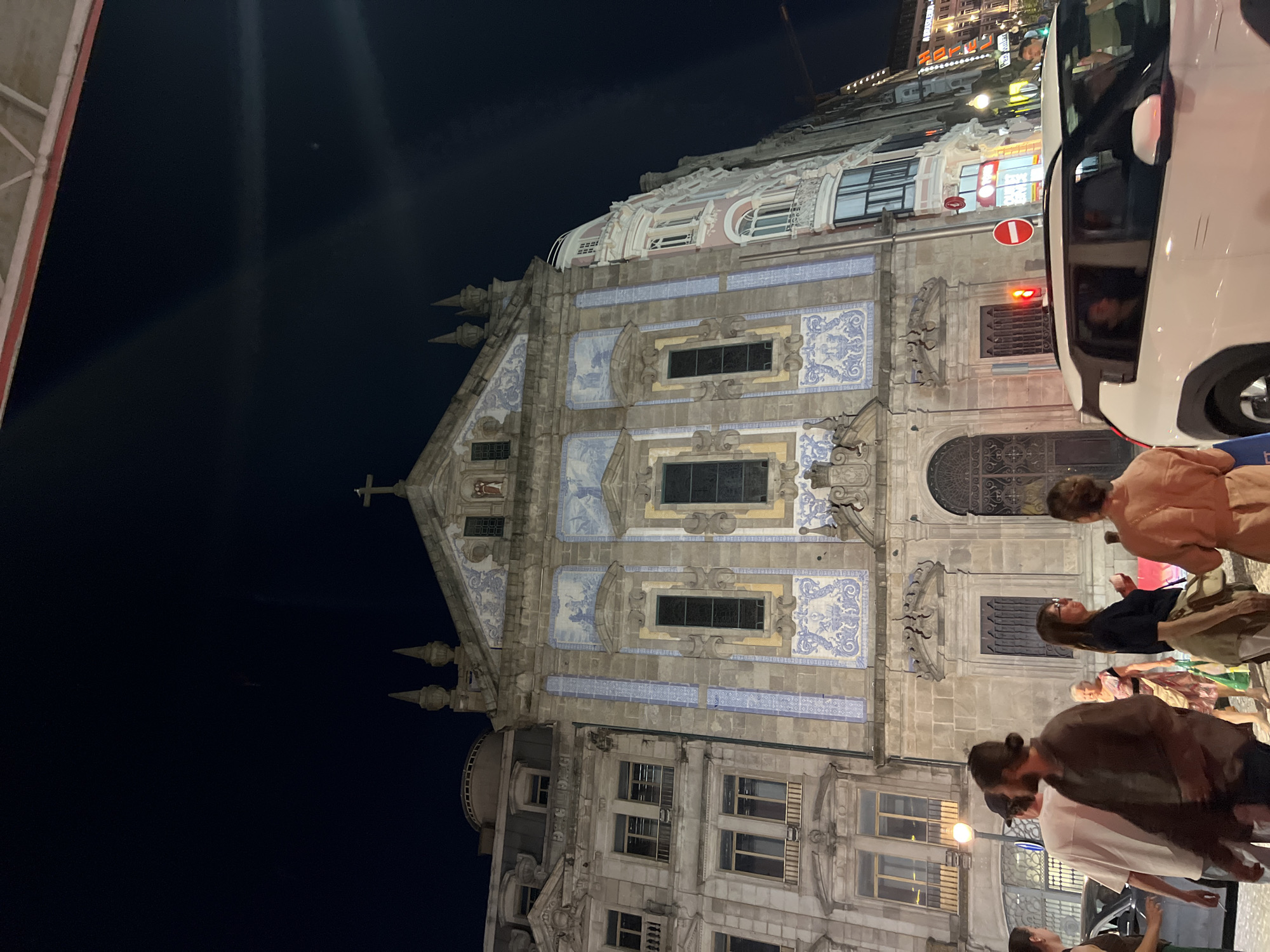 The image shows the Church of Saint Ildefonso in Vila Nova de Gaia, Portugal at night. The church facade is adorned with traditional blue and white azulejo tiles. In front of the church, several people are standing or walking, some interacting with each other. A white car is passing by on the street in the foreground. The street is illuminated by streetlights and light from buildings, casting shadows. The sky is dark, indicating nighttime. The architecture of the surrounding buildings suggests an urban setting.