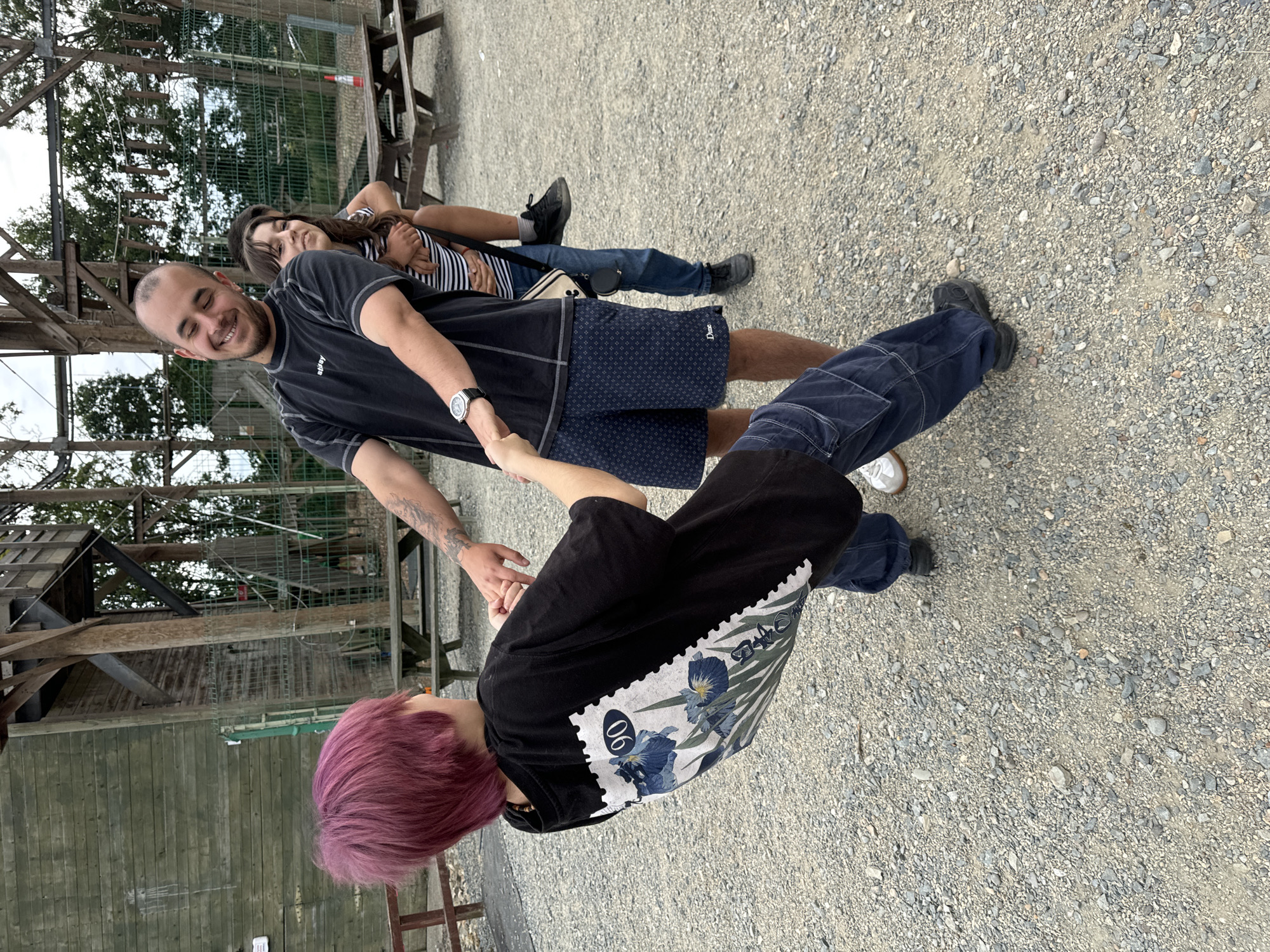 Three individuals are interacting outdoors on a gravel surface in front of a wooden structure. The person on the left, with short, vibrant pink hair, is facing away from the camera and is being pulled by the person in the center. They are wearing a black t-shirt with a graphic print on the back and blue cargo pants. The person in the center, a male with a beard, is smiling and holding the hands of the person on the left. He is wearing a black t-shirt and navy patterned shorts. Behind him, a person with dark hair is riding on his back. This person is wearing a striped t-shirt and jeans. In the background, there is a wooden structure, possibly part of an adventure park or playground, with fencing and trees. Picnic tables are visible further in the background. The lighting suggests it is daytime. This scene is located at Courtown Adventure Centre in Courtown, Ireland.