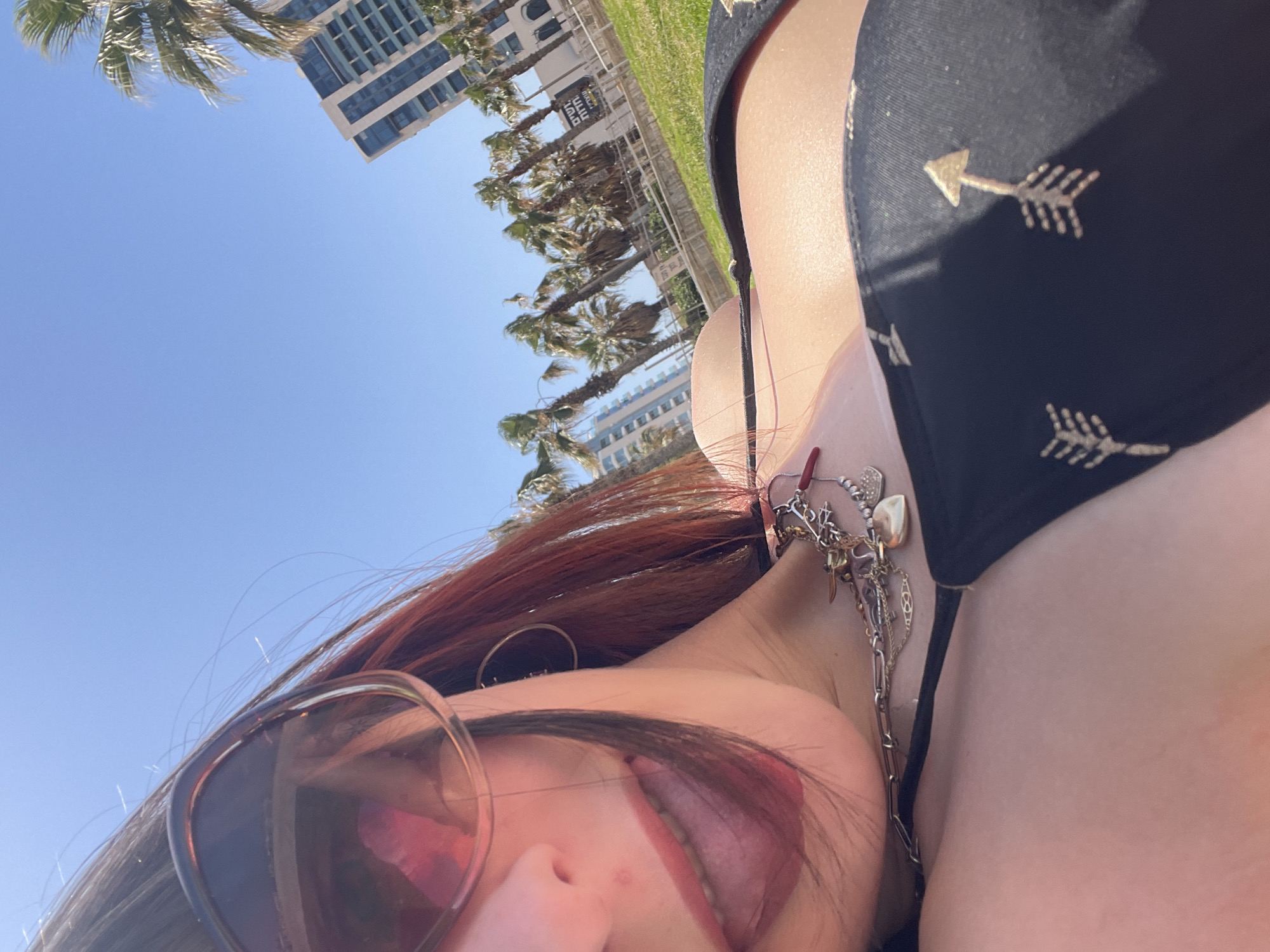 A selfie shows a woman with long auburn hair and sunglasses lying on her back, with her tongue sticking out, under a clear blue sky in Herzliya, Israel. In the background, palm trees line a green grassy area, and a modern high-rise building is visible. The woman is wearing a black bikini top adorned with gold arrow designs, and a layered necklace with various pendants.