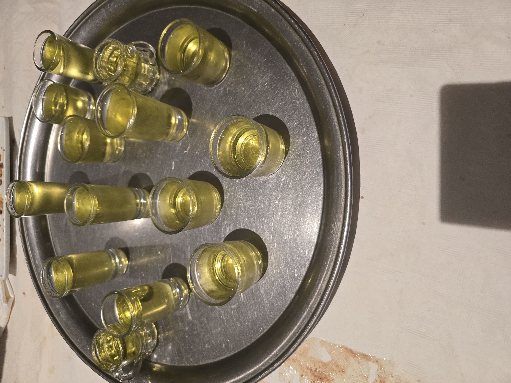 This image, taken in L'hospitalet De Llobregat, Spain, presents an overhead view of numerous small glasses, likely shot glasses, filled with a bright yellow liquid. The glasses are arranged on a large, circular, metallic serving tray that appears worn with scratches and scuff marks. There are at least 15 glasses visible, some clustered together, indicating a preparation for serving or a recent gathering. The liquid's color suggests it could be a liqueur, such as limoncello, or another type of shot.
The tray and glasses are positioned on a light-colored surface, possibly a tablecloth or a countertop, which shows some minor stains or smudges in the lower right portion. A distinct, dark, rectangular shadow is cast in the upper right corner of the frame, hinting at an artificial light source overhead. The overall lighting suggests an indoor setting, likely during the evening or night, given the contrast and shadow. No people are visible, but the arrangement of the shots implies a social event or celebration is taking place or has just occurred. There is no visible text in the image.