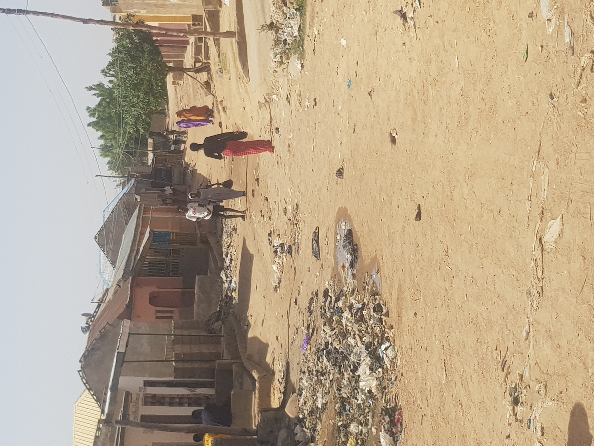 The image depicts an unpaved street in Dabawa, Nigeria, with visible litter scattered across the dirt road. Several individuals are present: two men are walking towards the viewer, while another person is seated near a building on the left side of the frame. Buildings with varying architectural styles line the street. Laundry is hung out to dry, and a tree is visible in the background.