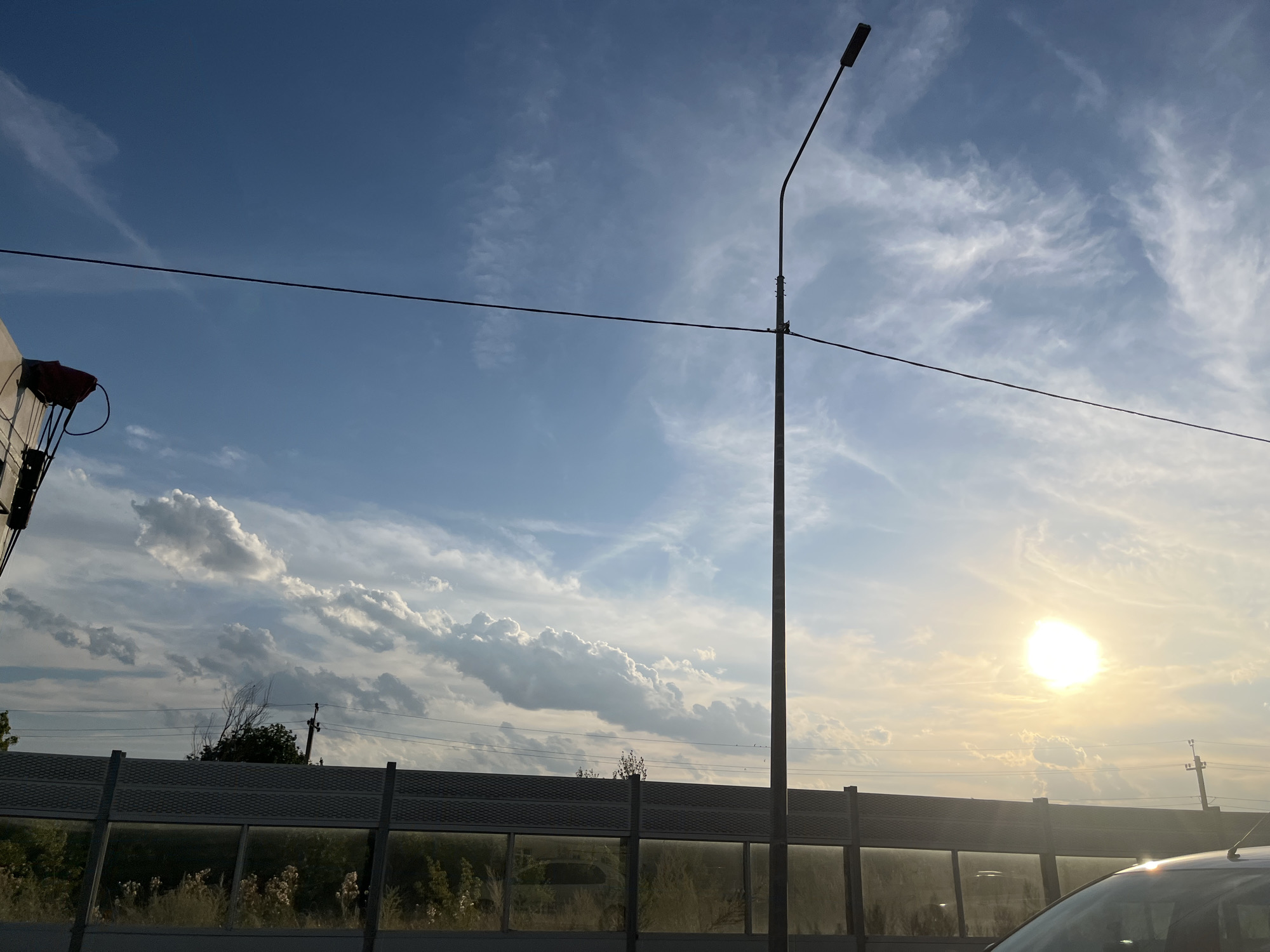 The image depicts a clear blue sky with scattered white clouds, illuminated by the bright sun setting on the right side of the frame. A tall, slender lamppost stands prominently in the center, with a dark horizontal wire stretching across the sky from the left edge. In the foreground, a sound barrier runs horizontally, constructed of dark gray material with reflective panels, behind which greenery and some trees are visible. Part of a white vehicle is visible in the lower right corner. The scene is consistent with an outdoor environment, possibly along a road or highway. While the location is specified as Пристанное, Russia, there are no specific visual cues within the image to confirm this particular location beyond the sky, lamppost, sound barrier, and vegetation. There are no people visible in the frame.