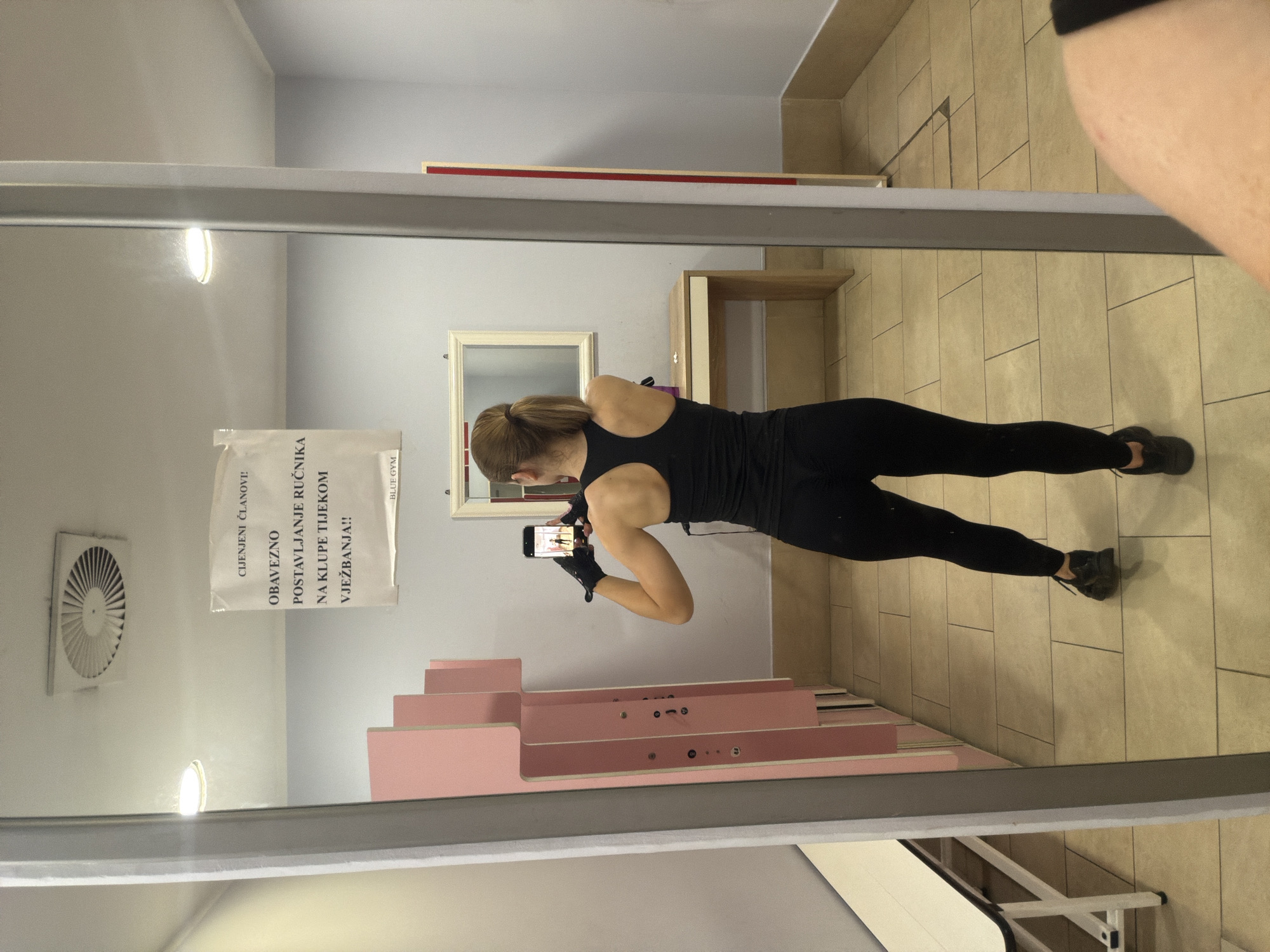 A woman is taking a mirror selfie in a gym locker room. She is wearing a black tank top, black leggings, black athletic gloves, and black sneakers. The locker room has pink lockers, a white bench, and tiled floors. There is a sign on the wall that reads "CIJENJENI ČLANOVI! OBAVEZNO POSTAVLJANJE RUČNIKA NA KLUPE TIJEKOM VJEŽBANJA!!". There is a mirror with a thin silver frame, and a woman is visible in the reflection, holding a cell phone and taking a photo.