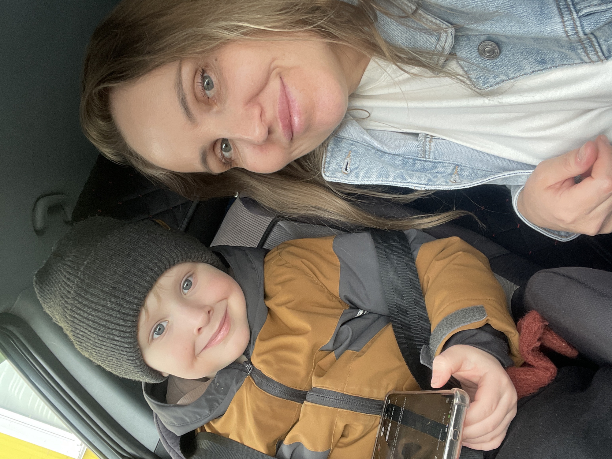 A selfie image depicts an adult female and a young child inside a vehicle. The adult female, with light brown hair and blue-green eyes, is positioned on the right side of the frame, smiling at the camera. She wears a light blue denim jacket over a white t-shirt. To her left is a young child, appearing male, with light blue eyes, wearing a dark grey knitted beanie and a brown and dark grey winter jacket. The child is secured by a black seatbelt and holds a smartphone in a clear case with his left hand, also smiling at the camera. A reddish-brown knitted item is visible near the child's lap. The vehicle's interior features dark grey upholstery. The background visible through the car window is out of focus, precluding the identification of a specific location within the city of Chaikovsky, Russia.