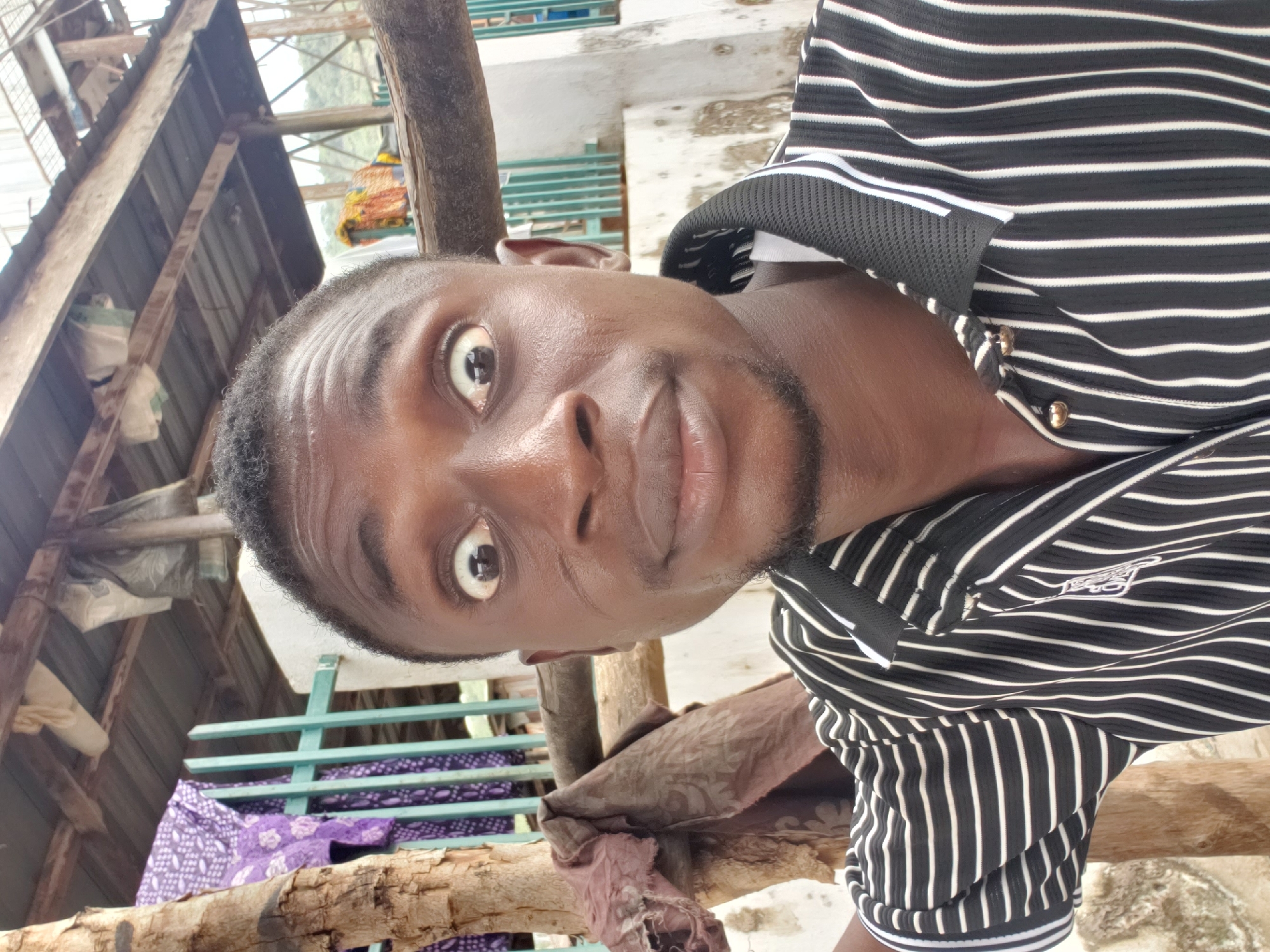 The image displays a close-up, tilted portrait of a young man with dark skin, short dark hair, and light facial stubble. He looks directly at the camera with wide, slightly surprised eyes and slightly parted lips, conveying a playful or mildly shocked expression. His skin appears somewhat shiny, indicating an outdoor setting or warm conditions.
He is wearing a black and white horizontal striped short-sleeved polo shirt. The collar features a black mesh-like material, and small gold-colored buttons adorn the placket. A small, indistinct white logo or symbol is visible on the left sleeve of his shirt.
The background suggests an outdoor or semi-outdoor setting in a rustic environment, consistent with Lokoja, Nigeria. Visible elements include rough wooden beams and posts, corrugated metal roofing, and white sacks or bags hanging. A vibrant, multi-colored decorative item, possibly a birdcage or woven lantern, is suspended in the background. Parts of a light blue-green slatted structure and a section of purple fabric with a white pattern, resembling traditional African print, are also discernible near wooden supports. The bright, even lighting indicates it is daytime.