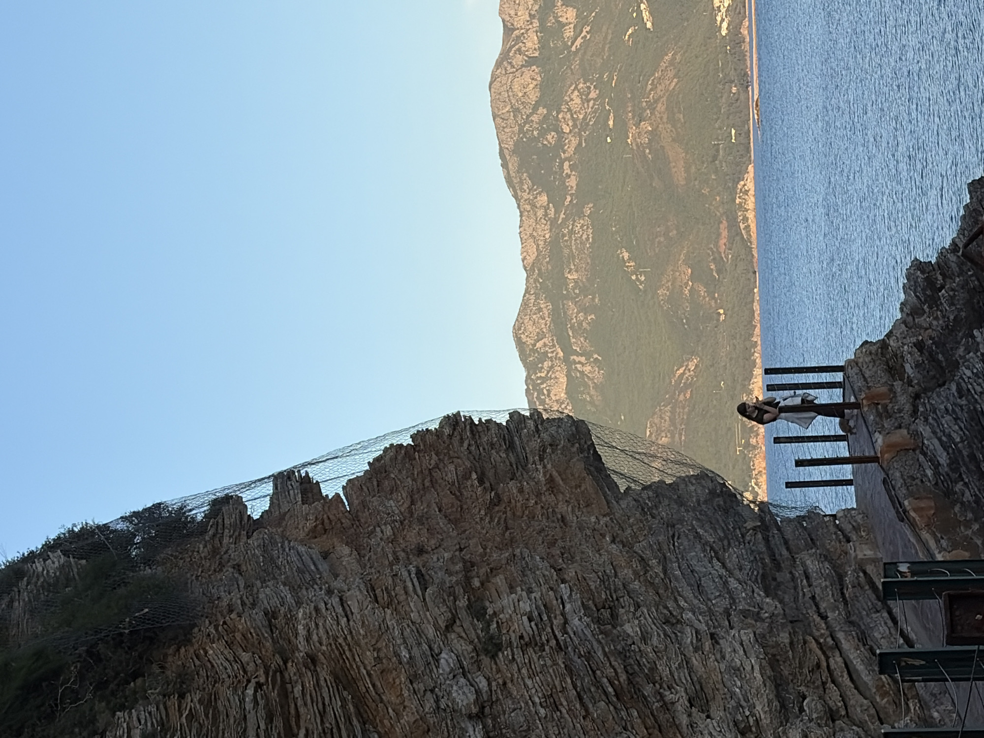 This image depicts a scenic coastal landscape in Budva, Montenegro, likely during late morning or late afternoon under a clear blue sky.
In the foreground, on the right side, a rugged and dark grey cliff face dominates the view, characterized by numerous vertical rock striations. Parts of this cliff are covered with a fine mesh netting, likely for rockfall protection. Integrated into the cliff are several human-made structures, including wooden planks and dark metal supports that form elevated platforms or a walkway.
On one of these platforms, positioned on the right side of the image, a lone individual, appearing to be a woman, stands. She is dressed in dark clothing, possibly with a light-colored top or jacket tied around her waist, and wears sunglasses. She is looking out towards the left, taking in the panoramic view.
Beyond the immediate cliff and platform, a vast expanse of calm, deep blue sea stretches across the mid-ground. Further in the distance, a range of large, imposing mountains, covered with some vegetation, rises across the water, forming the background horizon. The sky above is a clear, light blue, without any visible clouds.
The scene conveys a sense of tranquility and awe at the natural beauty of the coastline, experienced from a structured viewpoint. There is no visible text in the image.