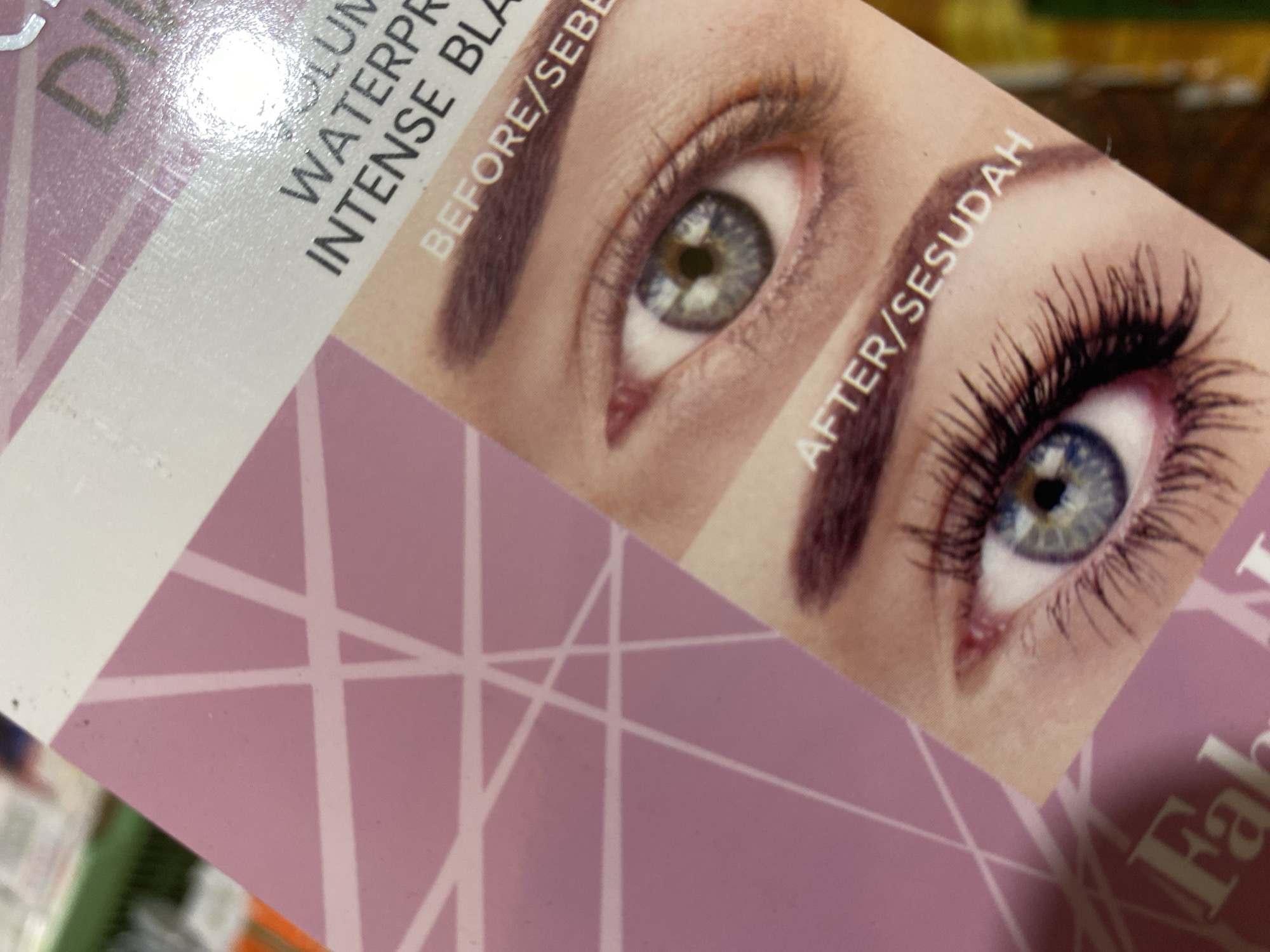 The image is a close-up of packaging for a mascara product, likely on display in a retail setting in Ta', Indonesia. The packaging features a split image of a woman's eye, showing a "before" and "after" effect of the mascara. Text on the packaging includes "DI VOLUM," "WATERPR," "INTENSE BLA," "BEFORE/SEBE," and "AFTER/SESUDAH." The background of the packaging is a light pink with white geometric lines. In the lower left corner, part of another product with the word "Fab" is visible. The overall impression is of a product advertisement for cosmetic enhancement.