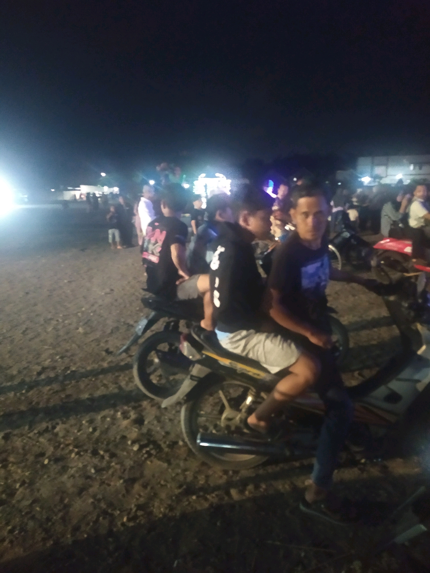In the city of Gempol, Indonesia, at night, two young men are sitting on a motorcycle on a dirt road, illuminated by artificial lights. Both are wearing dark t-shirts and shorts. The man on the right has a blue graphic on his t-shirt. The man on the left has a white graphic on his t-shirt. The background is filled with a crowd of people, many of whom are blurred and indistinct due to the low light and movement. Several motorcycles are visible throughout the crowd. The environment appears to be an open area, possibly a field or market, with makeshift structures and tents in the distance. The lighting suggests a festive or event-like atmosphere.