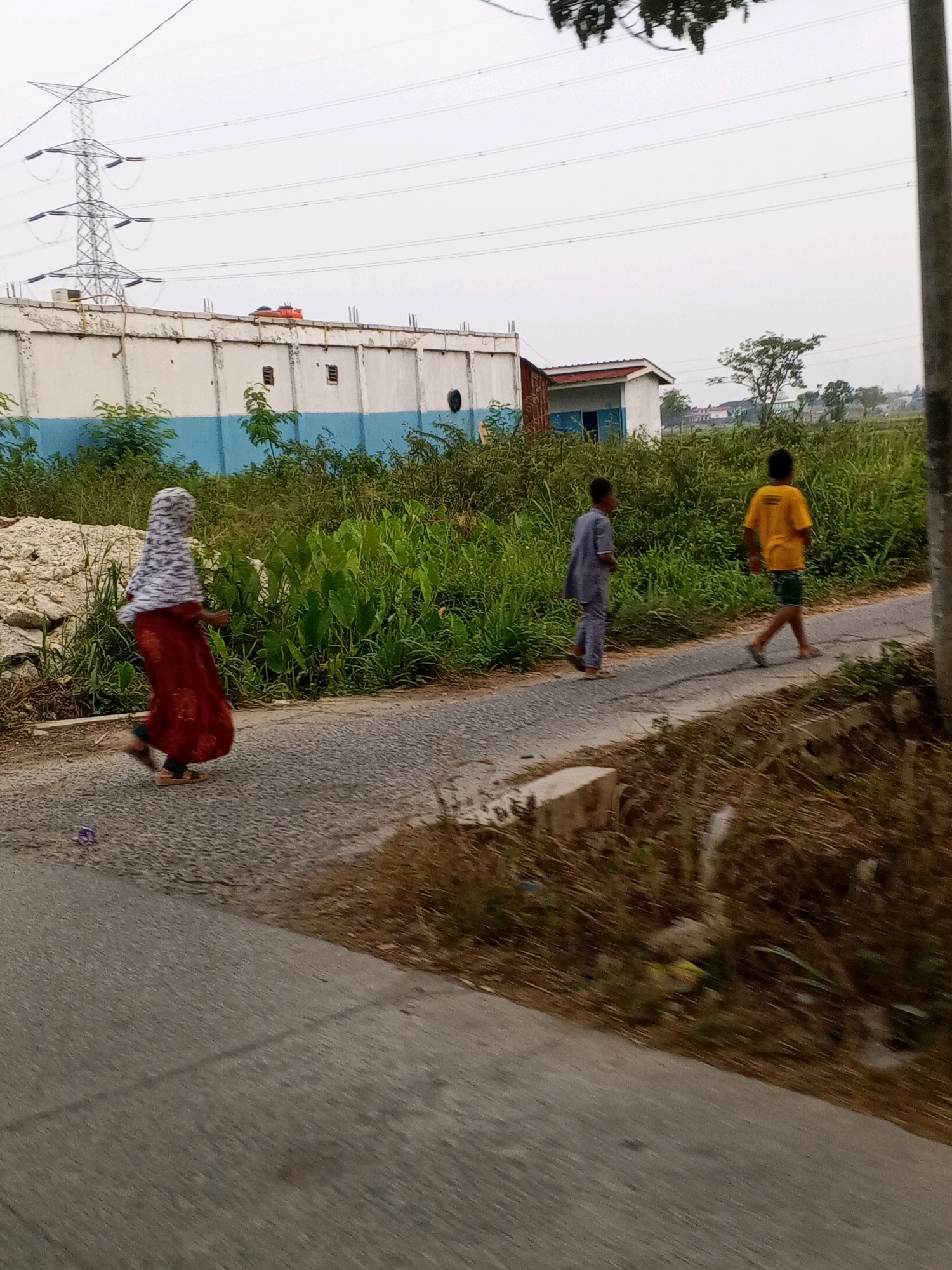 The image shows a view of a paved road in Ciruas, Indonesia, with a person in a red dress and headscarf running on the left side. Two boys are walking on the road ahead of the runner. The boy on the left wears a light blue garment, and the boy on the right wears a yellow t-shirt and green shorts. The road is bordered by dense green vegetation. In the background, there is a long, white building with a blue stripe near its base, and beyond that, a large metal pylon and overhead power lines. The sky is overcast. The scene appears to be in a semi-urban or rural area, likely during daylight hours.