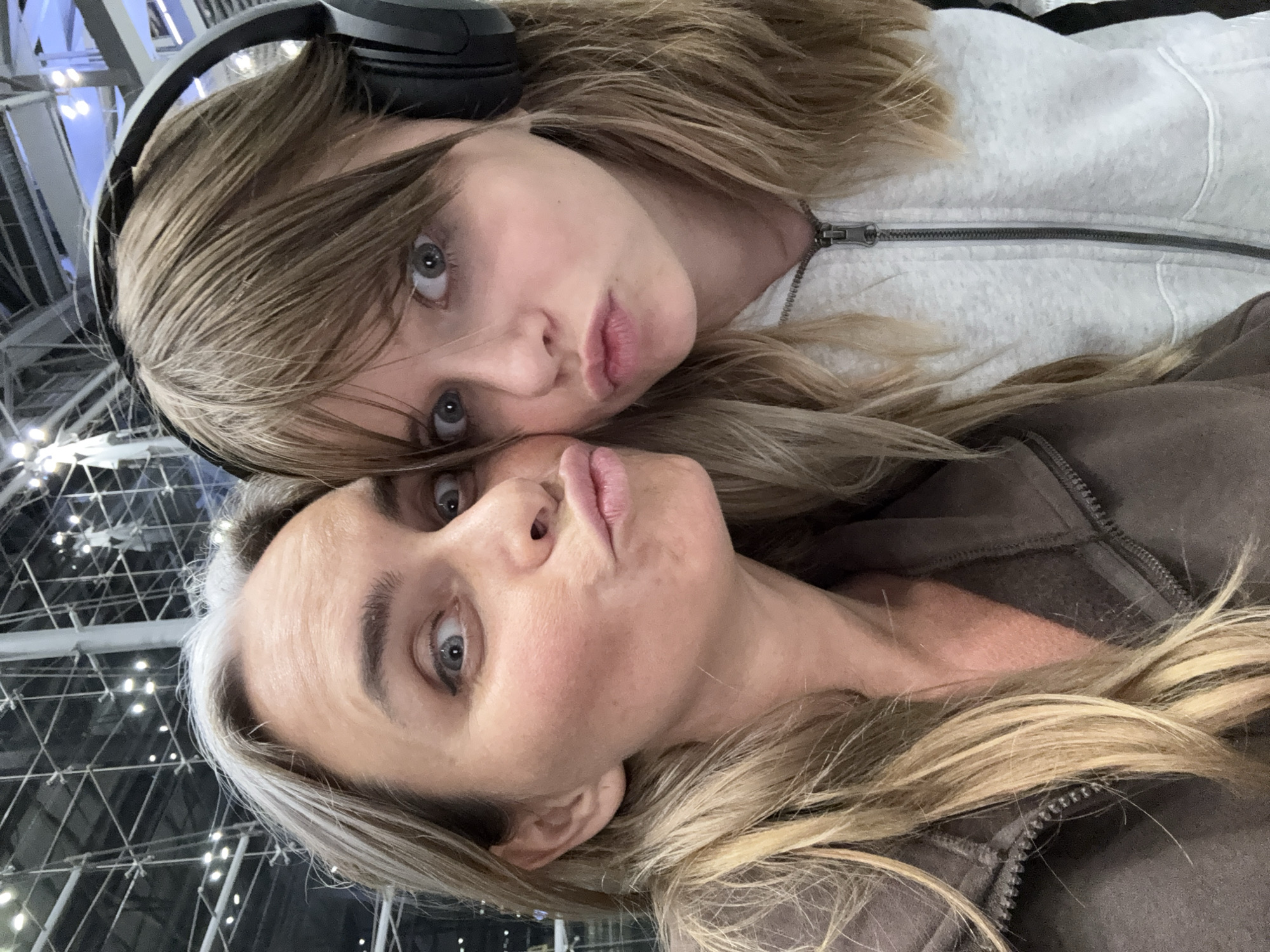 The image shows a close-up selfie of two females, an adult and a younger female, both with pouting expressions and wide eyes. They are positioned side-by-side, with the adult on the left and the younger female on the right. The adult has blonde hair with streaks of gray and is wearing a brown zip-up hoodie. The younger female has blonde hair and is wearing a light gray zip-up hoodie and headphones. The background appears to be an indoor setting with a complex structure of metal beams and lights, possibly a convention center or a large exhibition hall. The image was taken on December 7, 2023. The location is specified as Nong Prue Subdistrict, Thailand.