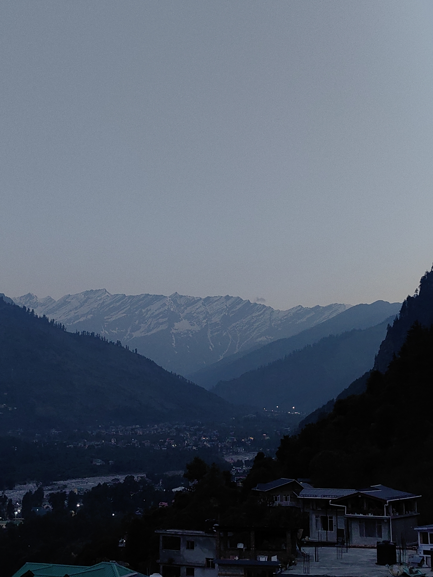 The image shows a twilight view of the valley in Vashisht, India. In the foreground, several modern buildings with flat roofs are visible. These buildings appear to be houses or guesthouses. A few are under construction, with exposed structural elements. In the middle ground, a small town or village is nestled in the valley, with numerous lights twinkling from houses and streets, indicating it is evening. Beyond the town, the valley slopes upwards, covered in dense, dark green pine trees. In the background, a majestic mountain range dominates the horizon. The peaks of these mountains are snow-capped and are illuminated by the fading light of dusk, with layers of hazy blue mountains visible in front of them, receding into the distance. The sky is a pale, hazy blue, characteristic of early evening or late twilight. There are no people or animals visible in the image.
