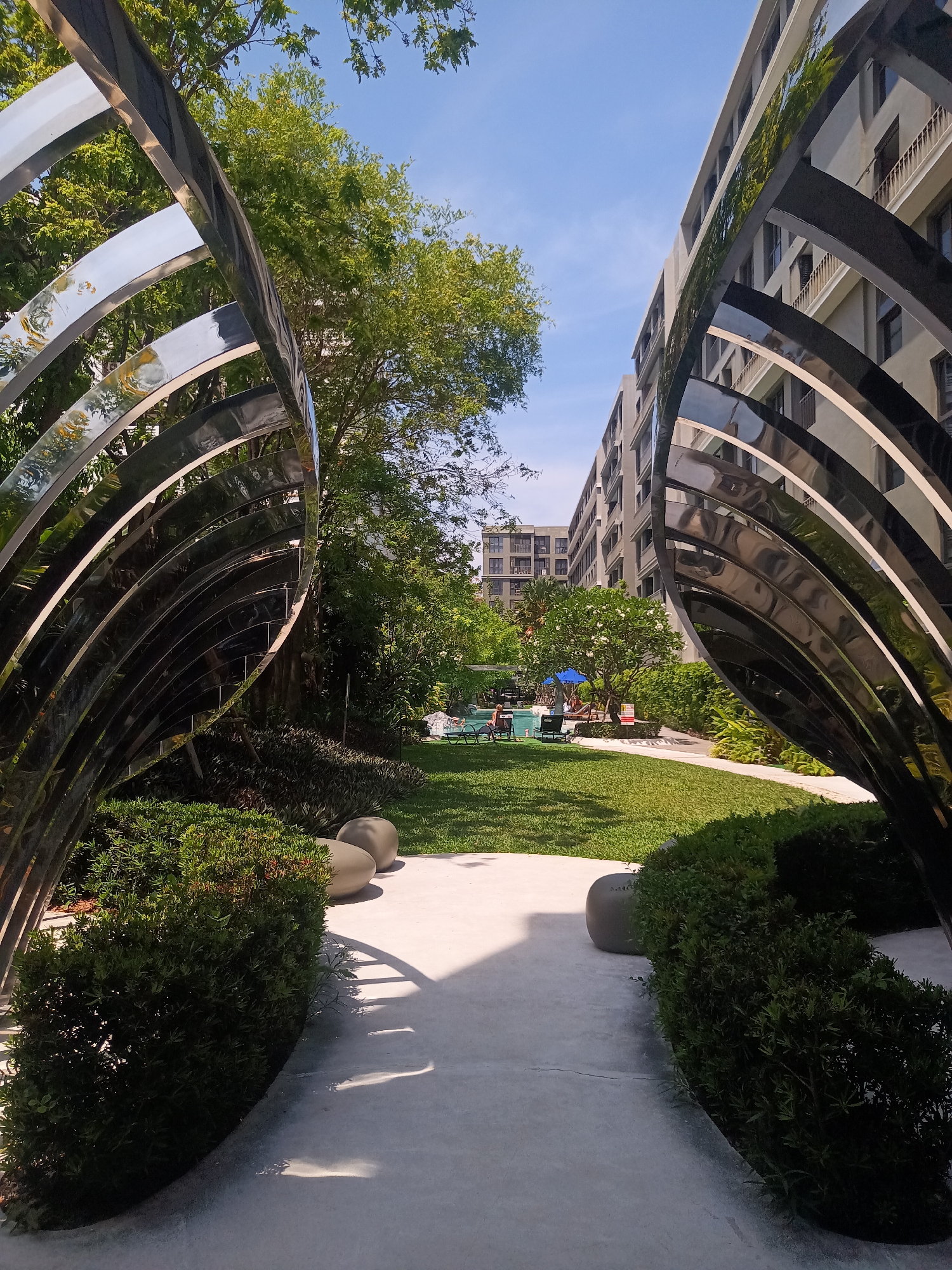 This image shows a manicured outdoor space in Hua Hin City Municipality, Thailand. The focal point is a pathway flanked by two curved, reflective metal structures resembling abstract wings or ribs. These structures frame a view of a green lawn, trees, and apartment buildings in the background.
The pathway is light-colored concrete, leading towards the center of the lawn. Green bushes and smooth, round stones are positioned near the base of the metal structures, adding to the landscaped design. Further down the lawn, a swimming pool area is visible, with blue umbrellas and lounge chairs indicating a recreational space.
The background shows multi-story apartment buildings, suggesting a residential complex. The sky is clear and blue, indicating a sunny day. The scene is generally tranquil and well-maintained.