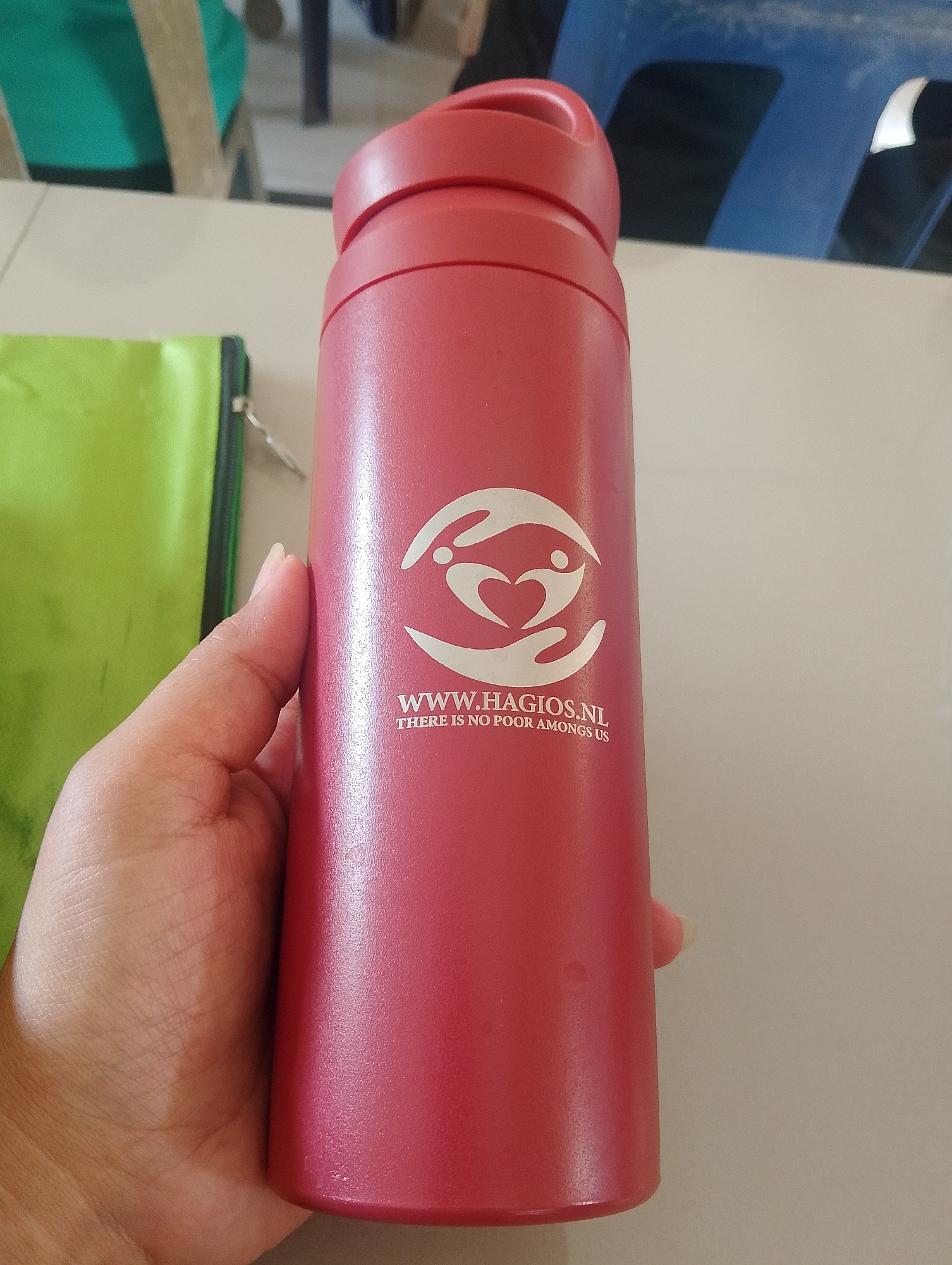 A close-up, eye-level, horizontal shot shows a hand holding a red reusable water bottle. The bottle has a white logo featuring two hands cupping a heart with a silhouette of a person inside. Below the logo, the text "WWW.HAGIOS.NL" is visible, followed by "THERE IS NO POOR AMONG US". A portion of a lime green bag with a black zipper is visible to the left of the bottle. The background is blurred, showing a light-colored table and hints of blue chairs and green fabric, suggesting an indoor setting. No specific location in Serbelawan, Indonesia can be identified from the image.