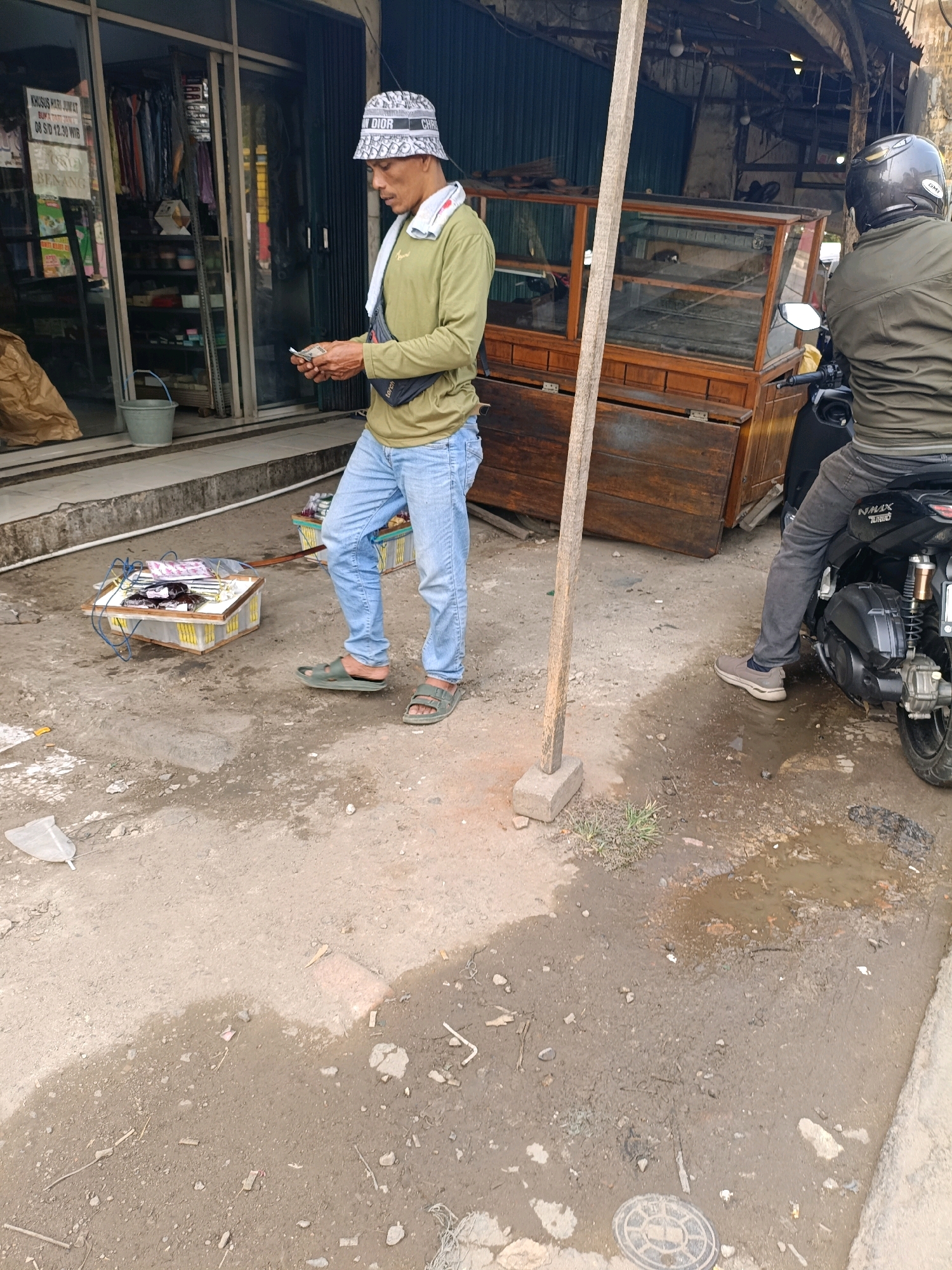 The image depicts an outdoor scene in Tangerang, Indonesia, on a dirt and paved ground surface. In the foreground, a man wearing a green long-sleeved shirt, blue jeans, and a bucket hat is holding money and a mobile phone. He has a towel draped around his neck and a black fanny pack. To his left, there is a small wooden structure with what appears to be merchandise, possibly cigarettes or small items, displayed in packaging and a plastic basket. In the background, a motorcycle rider wearing a dark green jacket and a black helmet is seated on a motorcycle, facing away from the camera. Behind them, there is a shop with a glass display case and wooden structures. The environment appears somewhat untidy with scattered debris and puddles of water on the ground, suggesting recent rain or a damp area.