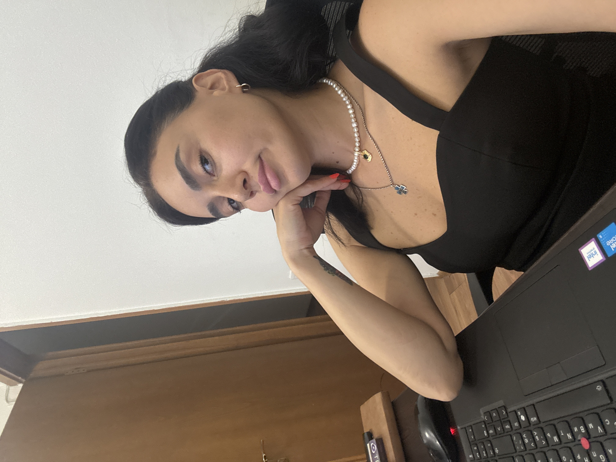 A young woman with dark hair and fair skin is sitting at a desk in a room. She is wearing a black sleeveless top and a pearl necklace with a gold pendant. Her right arm is resting on her chin, and she is looking at the camera. A laptop and a computer mouse are on the desk in front of her. A wooden door is visible in the background. The environment suggests an indoor setting, possibly an office or a home. There are no specific landmarks or indications that place the scene in Tbilisi, Georgia.