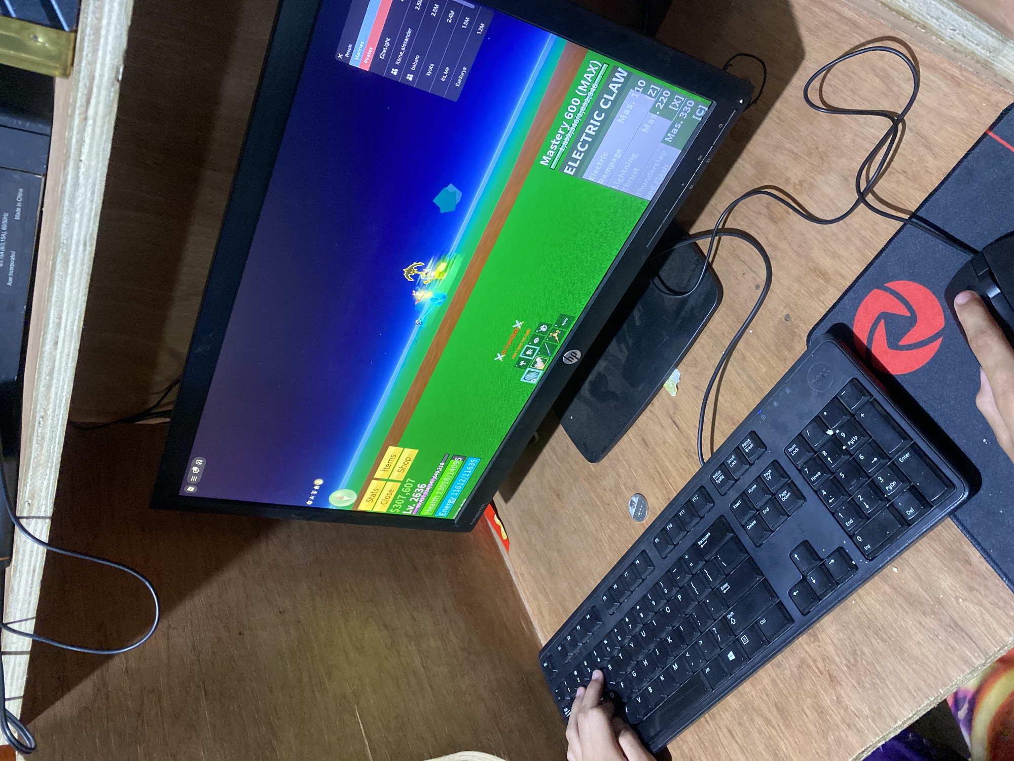 This image depicts a computer gaming setup located in Davao City, Philippines. The scene is an interior shot, showing a wooden desk and paneling.
In the foreground, a black Dell full-size keyboard is positioned on a light brown wooden desk surface. A child's or small adult's left hand rests on the left side of the keyboard, specifically near the 'W' and 'A' keys. To the right of the keyboard, a black computer mouse rests on a black mousepad featuring a red circular logo. A child's or small adult's right hand grips the mouse.
Central to the image is an HP monitor displaying a video game, identified by its user interface as "Blox Fruits" from Roblox. The game screen shows a vibrant green ground plane beneath a deep blue sky. Several in-game characters and visual effects are visible, including a character with yellow glowing elements. The game's heads-up display indicates a character level of 2636, current money at $307,607, health at 13016/14095, and energy at 11612/11635. "ELECTRIC CLAW" mastery is listed as 600 (MAX), with abilities mapped to 'Z', 'X', and 'C'. A player list is visible on the right side of the screen, showing usernames and associated values.
On the wooden desk, between the keyboard and the monitor, a single silver-colored coin (appearing to be a Philippine peso) is present. A small red and yellow object is tucked into the corner where the monitor meets the wooden wall.
Above the desk, a wooden shelf holds electronic components, including one marked "Acer incorporated" and "Made in China." A black cable hangs in a loop from this shelf.