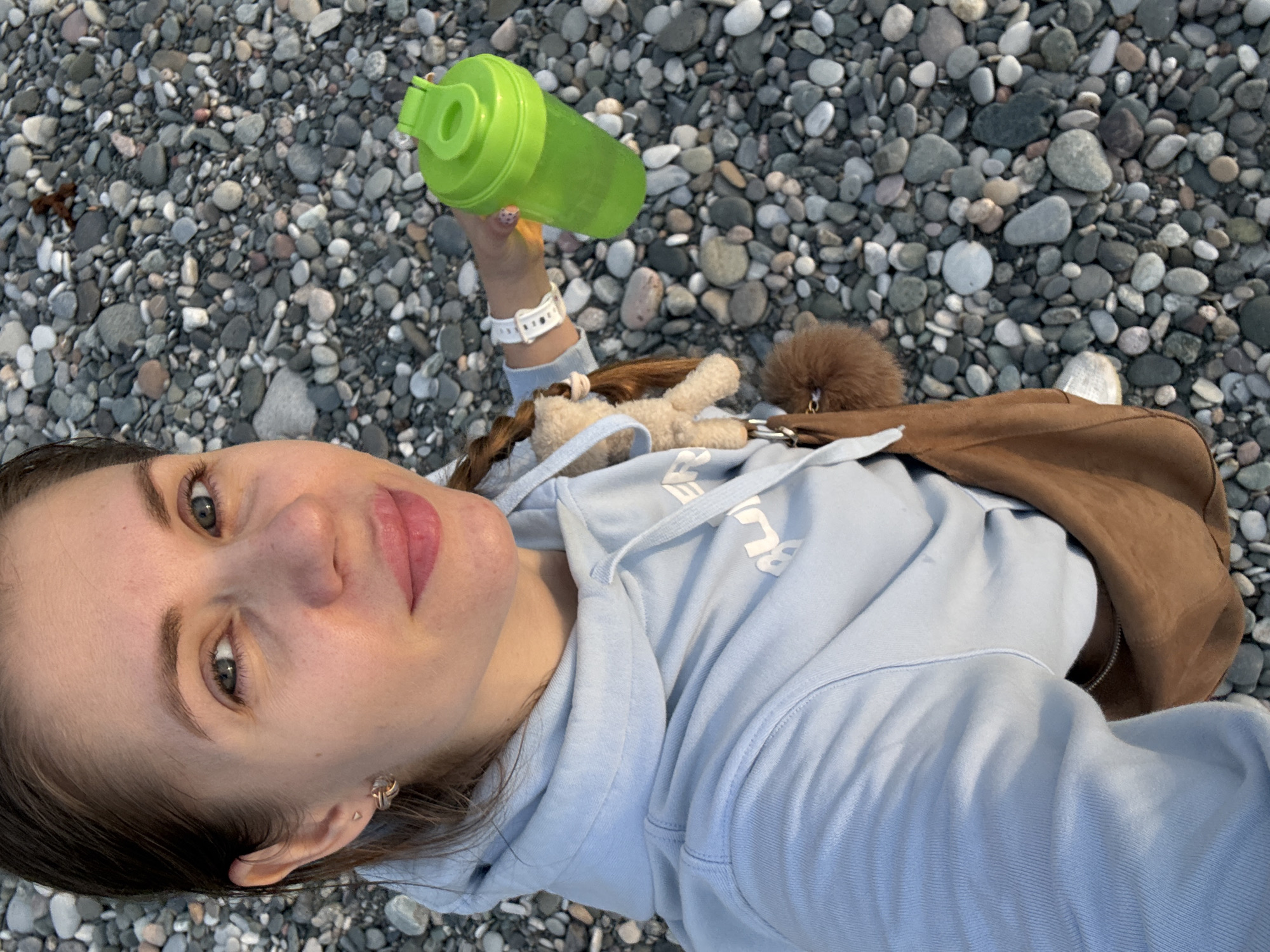 A woman lies on a beach composed of small pebbles of varying sizes and shades of gray, white, and black. She wears a light blue hooded sweatshirt and a brown crossbody bag. She holds a green water bottle in her right hand. A white smartwatch adorns her wrist. A plush toy bunny and a fluffy brown pom-pom are visible next to her shoulder. The location is likely a beach in Batumi, Georgia, judging by the pebble composition and the overall feel of the image.