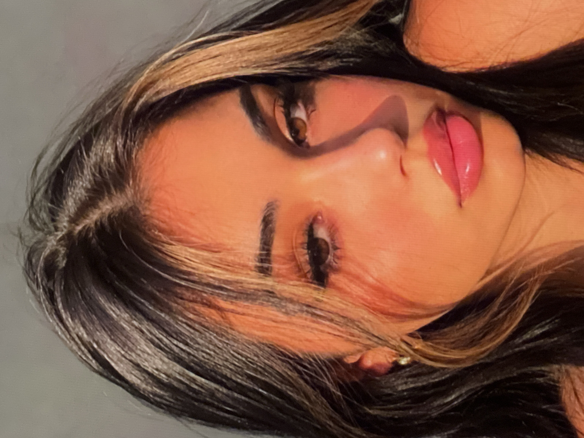 A close-up portrait of a young woman with dark hair and prominent makeup. She is looking slightly off-camera, with her head tilted. Her lips are full and painted a bright pink gloss. She wears dark eyeliner and mascara, and her eyebrows are well-defined. A single strand of blonde-highlighted hair falls across her forehead. The lighting suggests a warm, possibly outdoor setting, with a strong shadow cast across her cheek. The background is a plain, muted gray. An earring is visible on her left ear. The image is in portrait orientation.