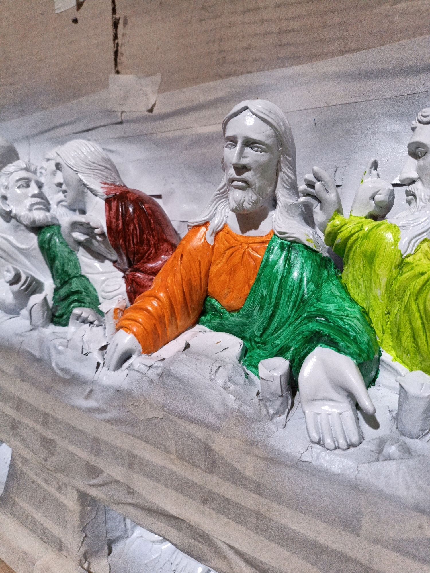 The image shows a close-up of a statue depicting the Last Supper, with some figures partially painted. The statue appears to be under a protective covering of paper or tarp. The figures are in white plaster or a similar material, with some areas painted in vibrant colors, including orange, green, and red. The figure of Jesus in the center is wearing a partially painted orange and green robe. Other figures have painted green or red garments. The statue is set against a textured, light-colored background, possibly a wall or ceiling, with some tape visible. There is no discernible information in the image that allows for the identification of its specific location in Cikal, Indonesia.