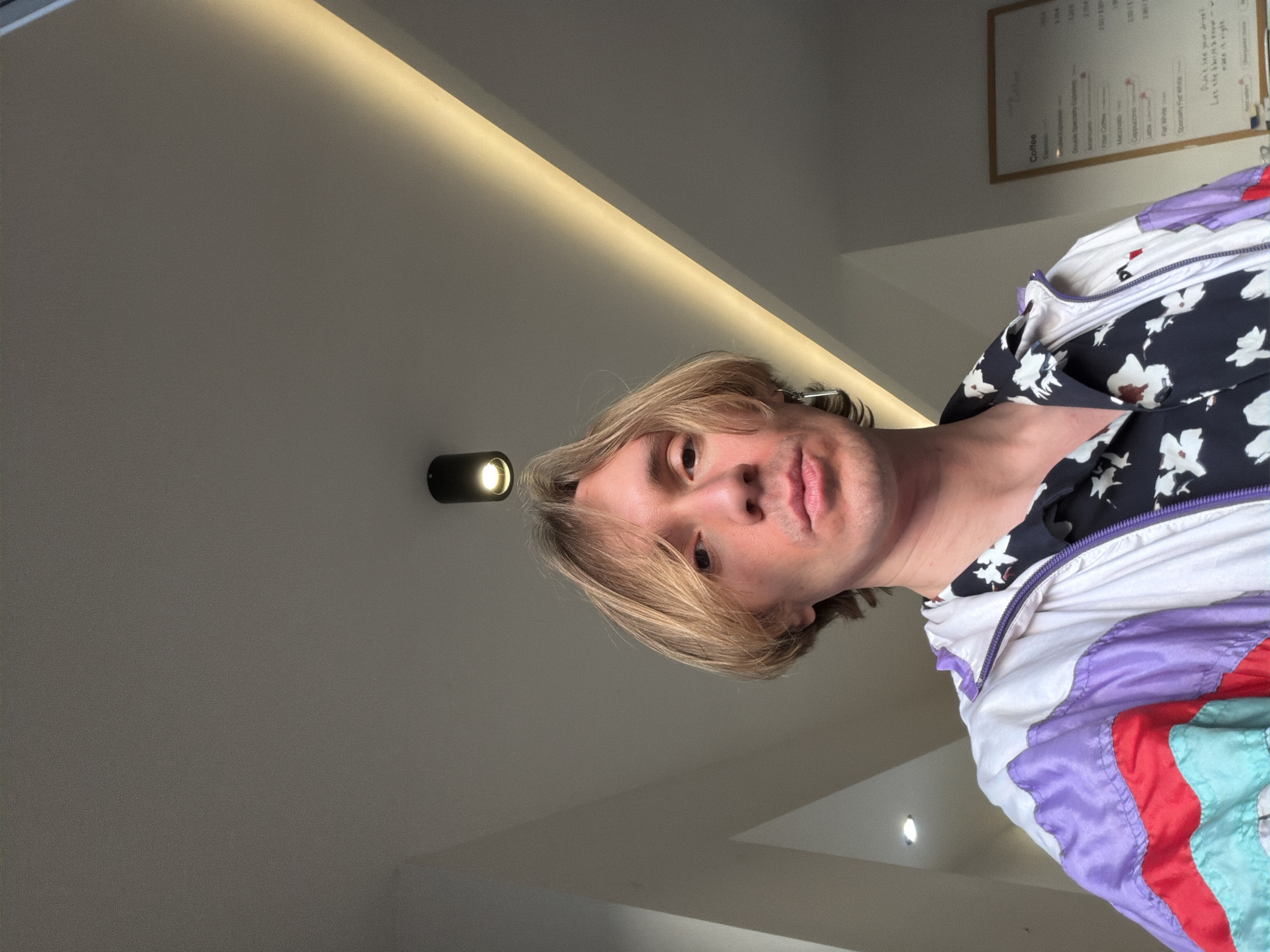 The image is an upward-facing selfie of a fair-skinned person with blond hair, wearing a floral-patterned shirt beneath a white jacket with purple, red, and turquoise stripes. A small, round, wall-mounted light fixture is visible in the background. To the upper right, a menu board with printed text detailing coffee prices is affixed to the wall. The photo appears to be taken indoors, possibly in a cafe, given the menu board's context. The precise location in Budva, Montenegro, cannot be determined from the visual information provided.