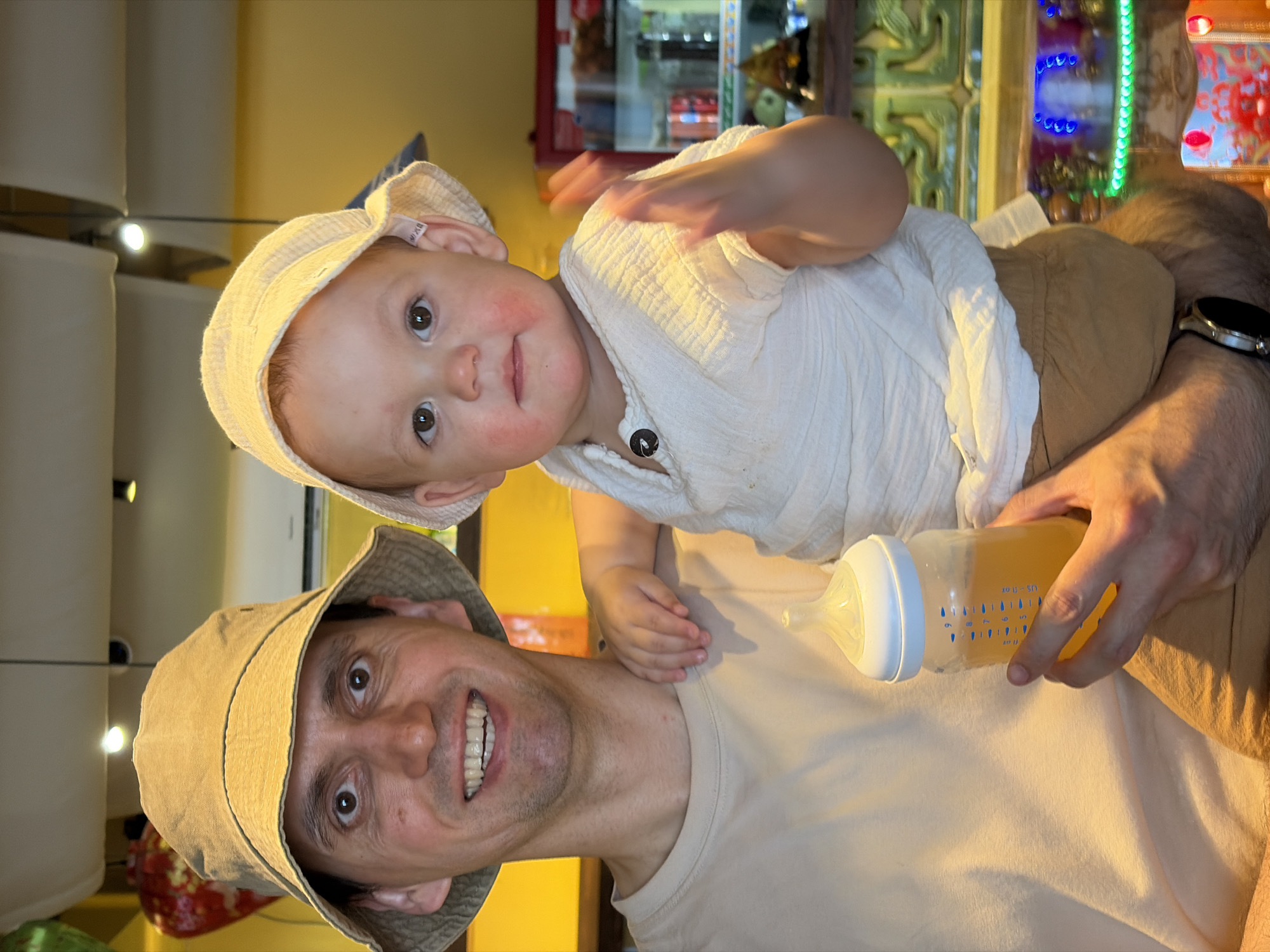 A man wearing a light-colored bucket hat is holding a baby in his arms. The baby is wearing a light-colored sun hat and a white shirt. The man is holding a baby bottle with a yellow liquid inside. The background appears to be indoors with shelves containing various items, possibly in a shop or restaurant. I cannot determine the specific location in Đà Nẵng, Vietnam based on the visual information provided.