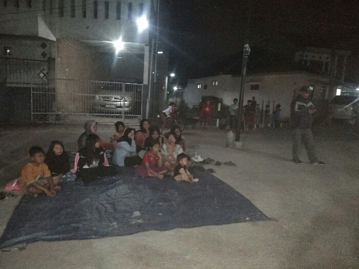 The image captures a nighttime scene in Baleendah, Indonesia, showing a group of people gathered outdoors on what appears to be a street or open paved area. The primary subjects are a diverse group of individuals, predominantly children and women, sitting close together on a large, dark blue tarp laid on the ground in the foreground. They are mostly facing towards the right side of the frame, some appearing to look off-camera, while others are interacting amongst themselves. Their expressions are generally subdued or attentive, though difficult to discern clearly in the low light.
To the right of this seated group, a man wearing a cap and a jacket stands, engrossed in his mobile phone. Further in the background, a small dog and several other individuals, including adults and children, are standing along the side of the road. The setting is illuminated by various artificial lights, including streetlights and lights from nearby houses, creating bright spots and deep shadows.
In the background to the left, a house with visible windows and a metal gate can be seen, behind which a silver car is parked. Other residential buildings are visible in the distance, further suggesting a neighborhood setting. The overall activity suggests a communal gathering, possibly for watching an event, seeking temporary refuge, or simply socializing in the evening. The time of day is clearly night, characterized by the darkness and reliance on artificial illumination. No visible text is clearly legible within the image.