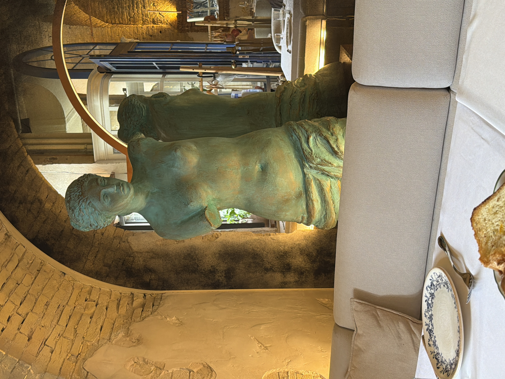 The image depicts an interior scene within an establishment in Kyiv, Ukraine. Prominently featured is a full-body statue of a woman, rendered in a verdigris patina, positioned in the foreground. The statue appears to be a classical or neoclassical style representation, with missing arms. Behind the statue, a mirrored surface reflects another statue, seemingly identical, creating a sense of depth. The background reveals an arched brick ceiling, typical of older architecture, and a large window or doorway with glass panes and dark framing, offering a glimpse into another part of the establishment. Lighting in the scene is warm and ambient, casting shadows and highlighting textures. In the immediate foreground, a white tablecloth covers a table, on which a decorative plate with blue floral patterns and a fork are visible. A slice of bread or cake is also partially visible at the bottom of the frame. The overall ambiance suggests a restaurant, cafe, or gallery setting, characterized by its architectural details and artistic display.